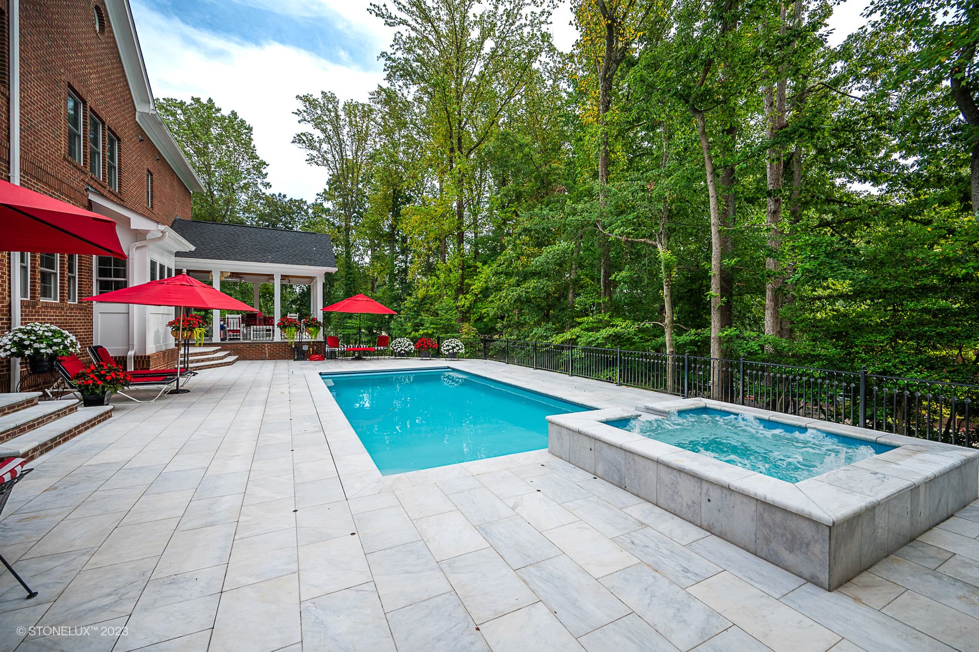 A backyard with a rectangular swimming pool and adjacent hot tub, surrounded by a light stone patio in White Whisper: The Pristine Afyon Marble Pool Elegance. Red umbrellas and lounge chairs sit near a brick house, framed by dense green trees.