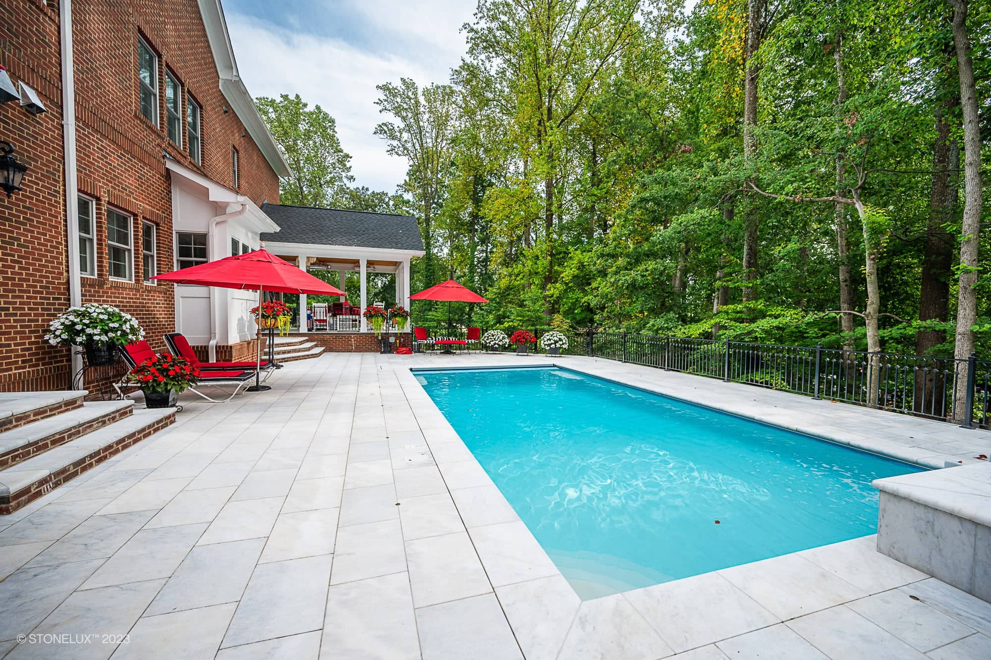 A rectangular swimming pool with clear blue water is surrounded by a tiled patio. Red umbrellas shade lounge chairs and tables, while the White Whisper: The Pristine Afyon Marble Pool Elegance adds a touch of luxury to the serene setting.