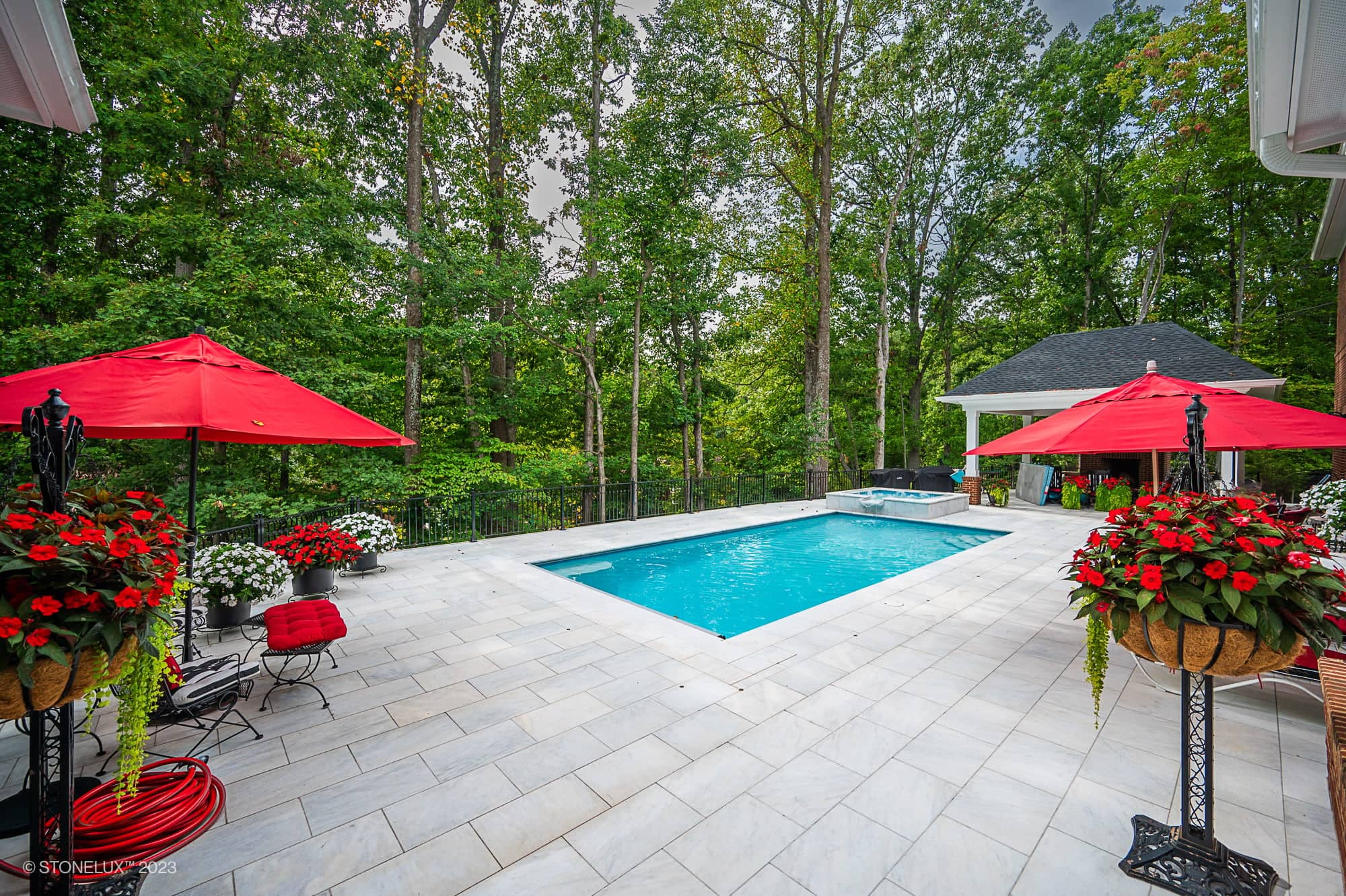 A rectangular backyard pool surrounded by White Whisper: The Pristine Afyon Marble Pool Elegance, white stone tiles, red umbrella-covered seating areas, and lush green trees. Potted red and white flowers decorate the patio beside a small pool house.