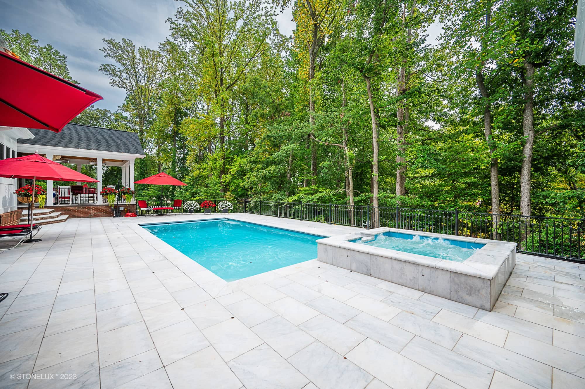 A spacious backyard features a rectangular swimming pool crafted with White Whisper: The Pristine Afyon Marble Pool Elegance, an adjacent hot tub, and a white stone patio with red umbrellas, lounge chairs, and lush green trees in the background.