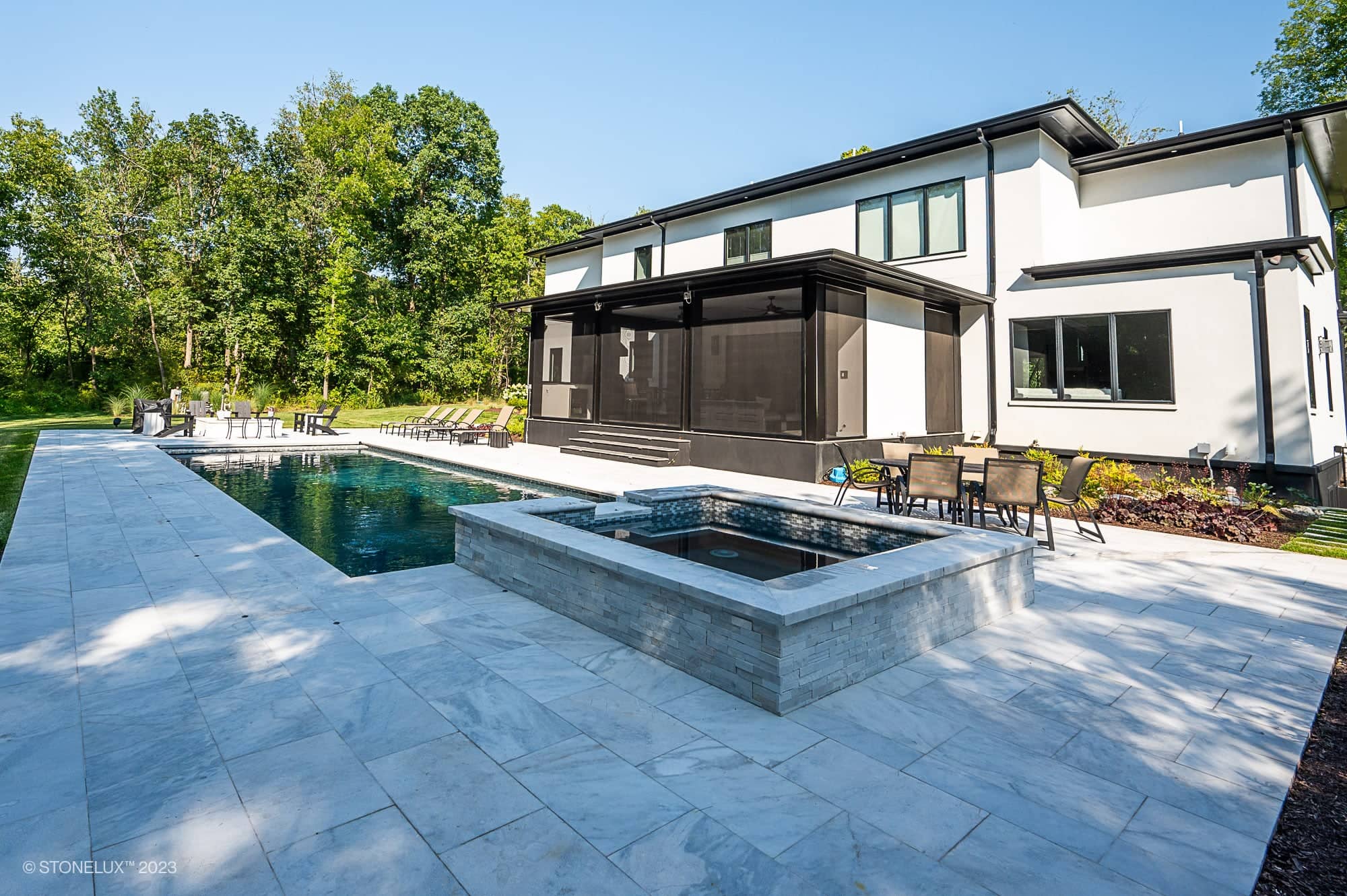 Modern backyard featuring a rectangular swimming pool and hot tub, with lounge chairs and patio tables. The Afyon Elegance: Modern White Marvel with Black Accents Poolside complements the two-story house surrounded by lush trees and greenery.