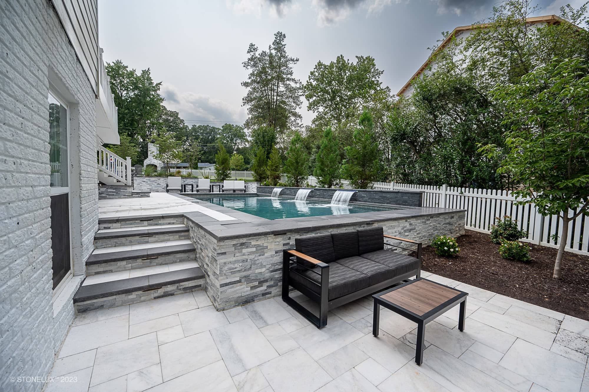 Modern backyard with a sleek in-ground pool featuring Modern Monochrome: The Afyon White Marble & Black Basalt Poolside Harmony, waterfall fountains, white fence, patio seating, stone steps, and lush greenery under a partly cloudy sky.