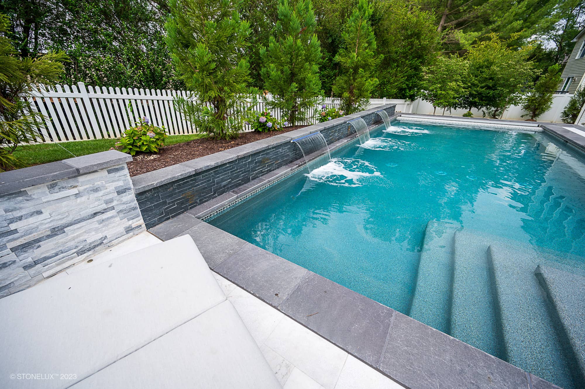 A modern backyard pool with Afyon White Marble and Black Basalt accents, small waterfalls along one edge, steps into the water, and a white picket fence framed by green trees—Modern Monochrome: The Poolside Harmony.