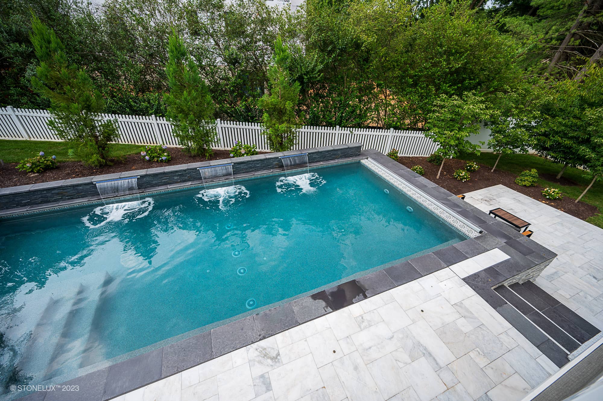 A rectangular backyard pool with water jets is framed by Modern Monochrome: The Afyon White Marble & Black Basalt Poolside Harmony, surrounded by stone tiles, lush greenery, a white picket fence, and a wooden bench on the stone patio.