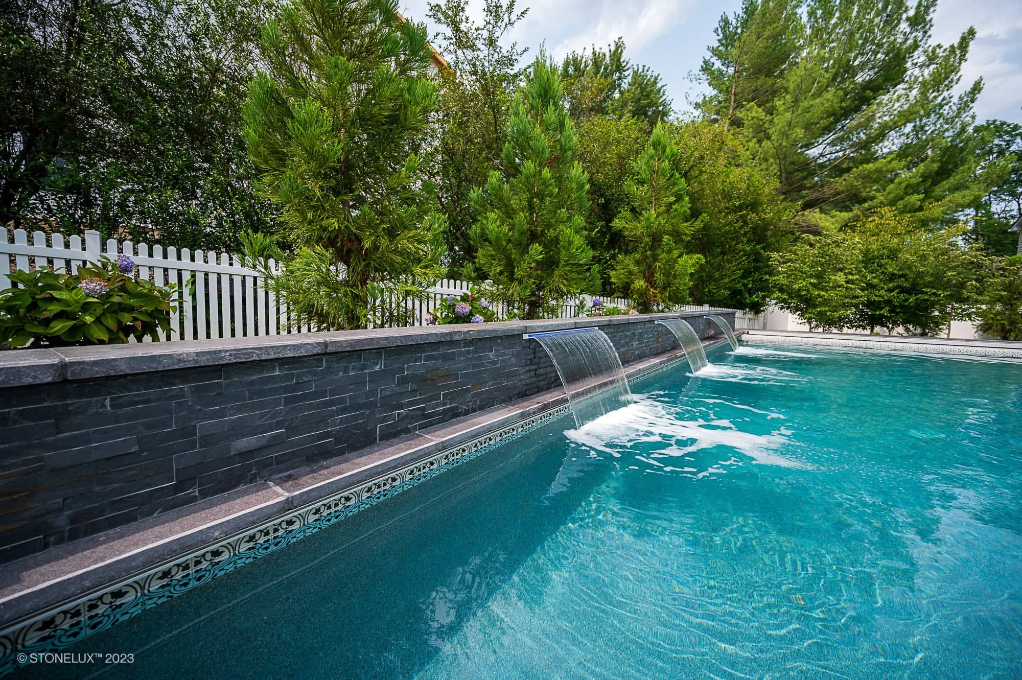 A clear backyard swimming pool features three small waterfall fountains flowing from a stone wall of Afyon White Marble & Black Basalt, surrounded by greenery, trees, and a white picket fence for modern monochrome harmony.