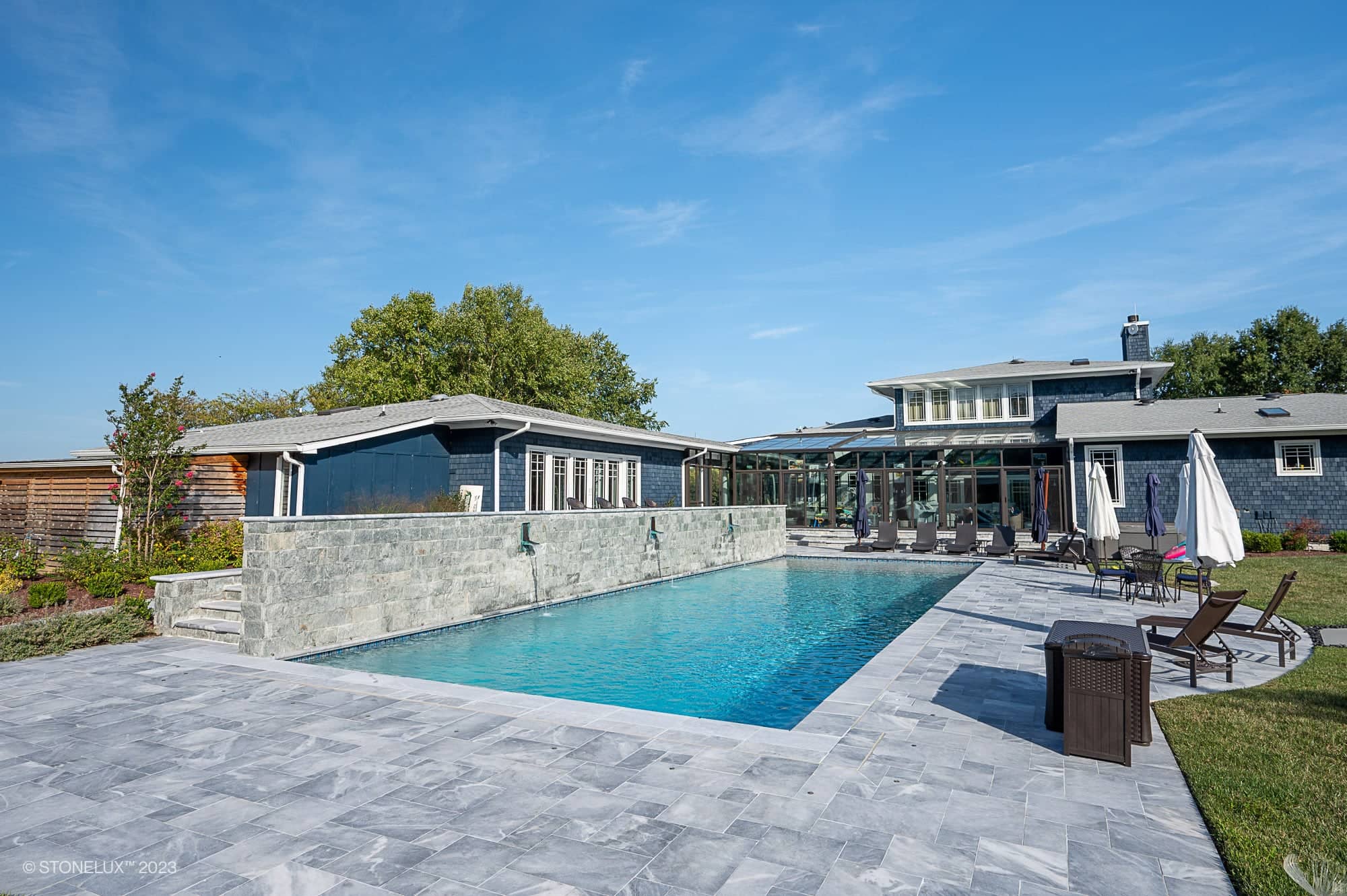 Modern backyard with a rectangular swimming pool—Blue Haven: The Grey and Blue Sandblasted Marble Pool Project—surrounded by gray stone tiles, lounge chairs, umbrellas, and a contemporary house in the background under a clear blue sky.