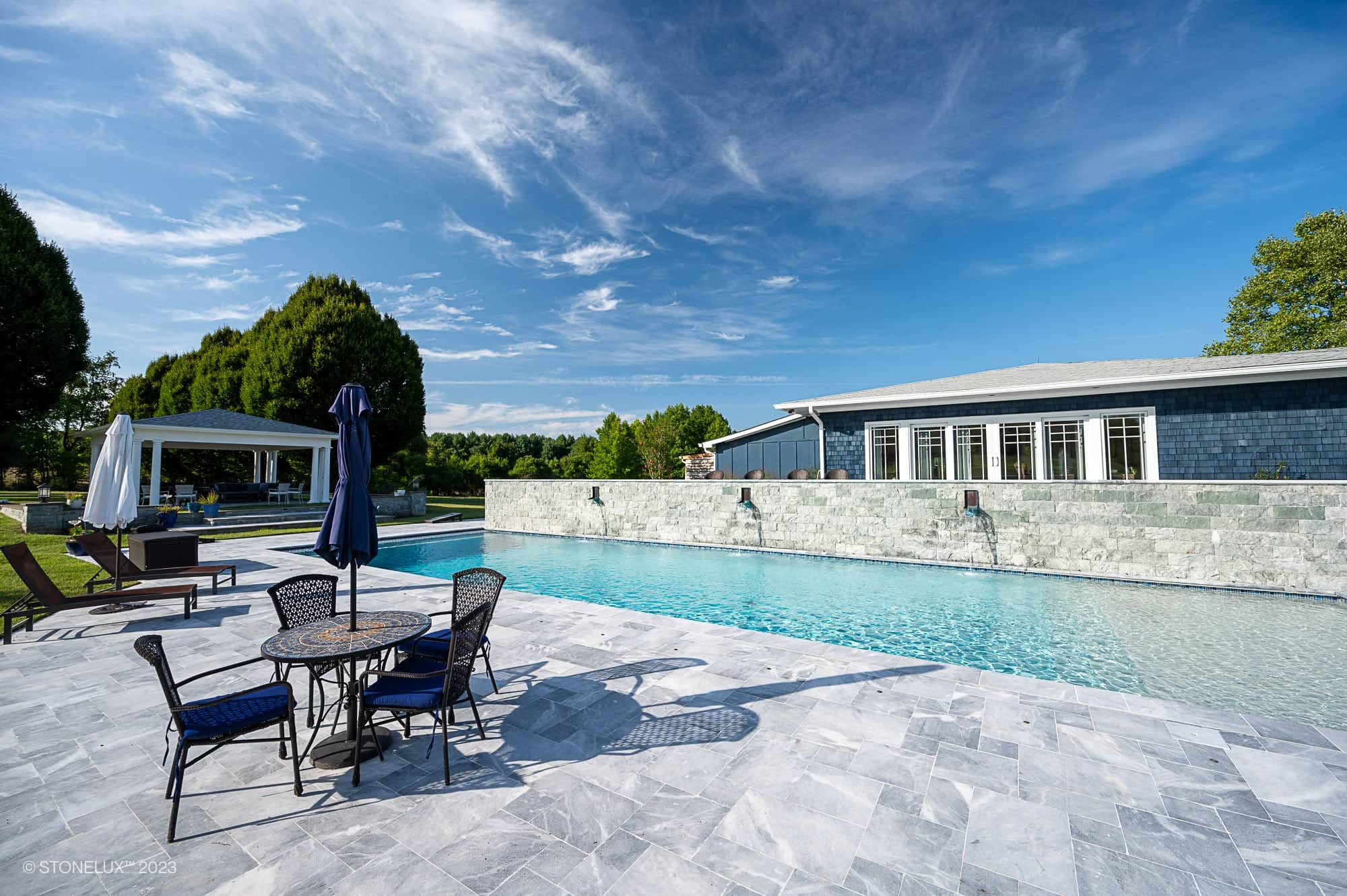 A sunlit outdoor pool area, part of the Blue Haven: The Grey and Blue Sandblasted Marble Pool Project, features a marble deck, round table with chairs under an umbrella, and a long swimming pool beside lush trees under a blue sky.