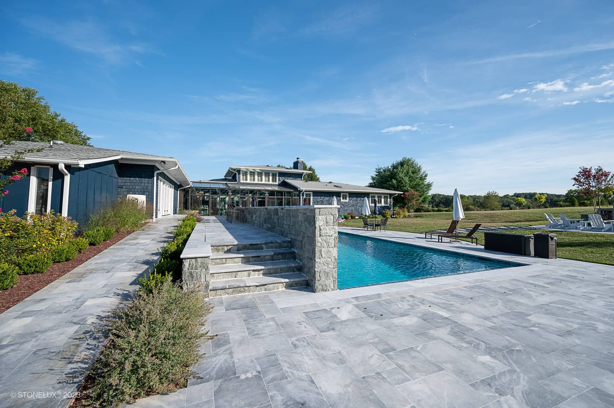 Modern outdoor swimming pool with stone tile deck, lounge chairs, and landscaped garden. Steps lead to Blue Haven: The Grey and Blue Sandblasted Marble Pool Project, with a contemporary house and large windows under a clear blue sky.