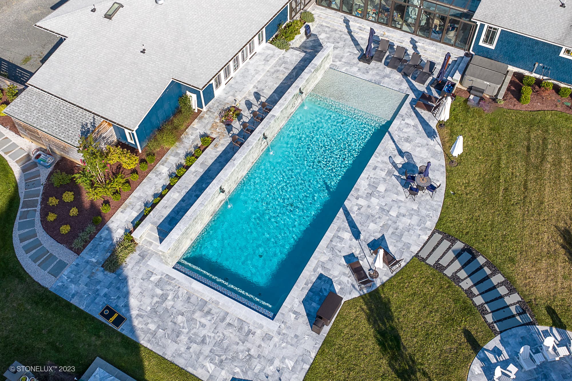 Aerial view of Blue Haven: The Grey and Blue Sandblasted Marble Pool Project, featuring a modern rectangular swimming pool with clear blue water, gray stone decking, lounge chairs, and landscaped grassy areas beside a building.