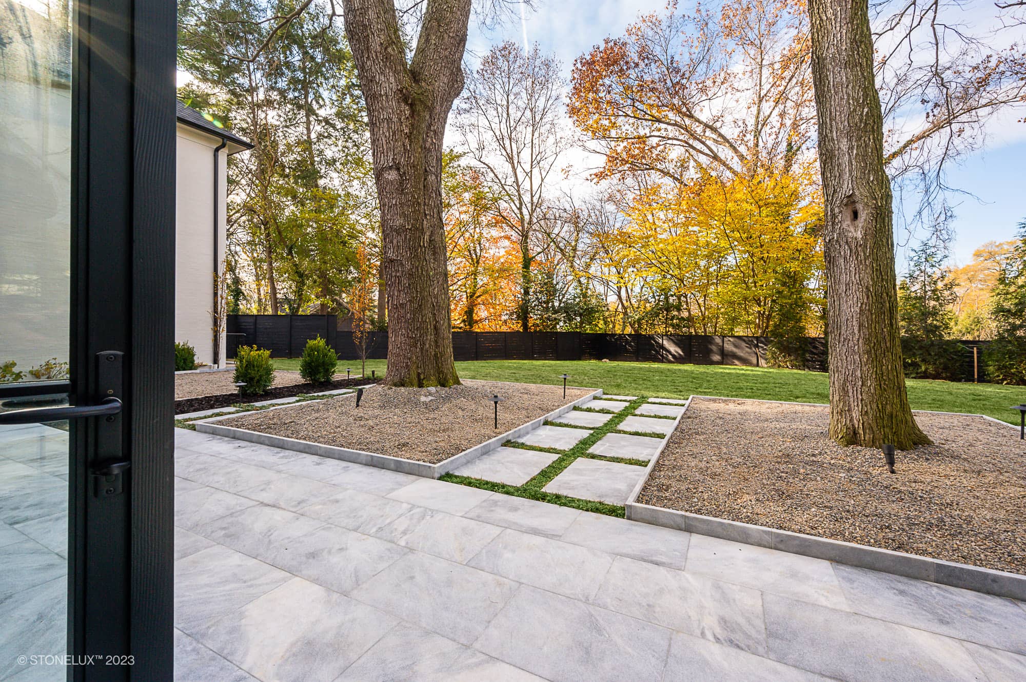 A modern backyard patio featuring Arctic Elegance: The Blue and Grey Sandblasted Marble Pool and Patio Project, with large stone tiles, gravel landscaping, two tall trees, stepping stones, and a black fence framed by autumn-colored trees.