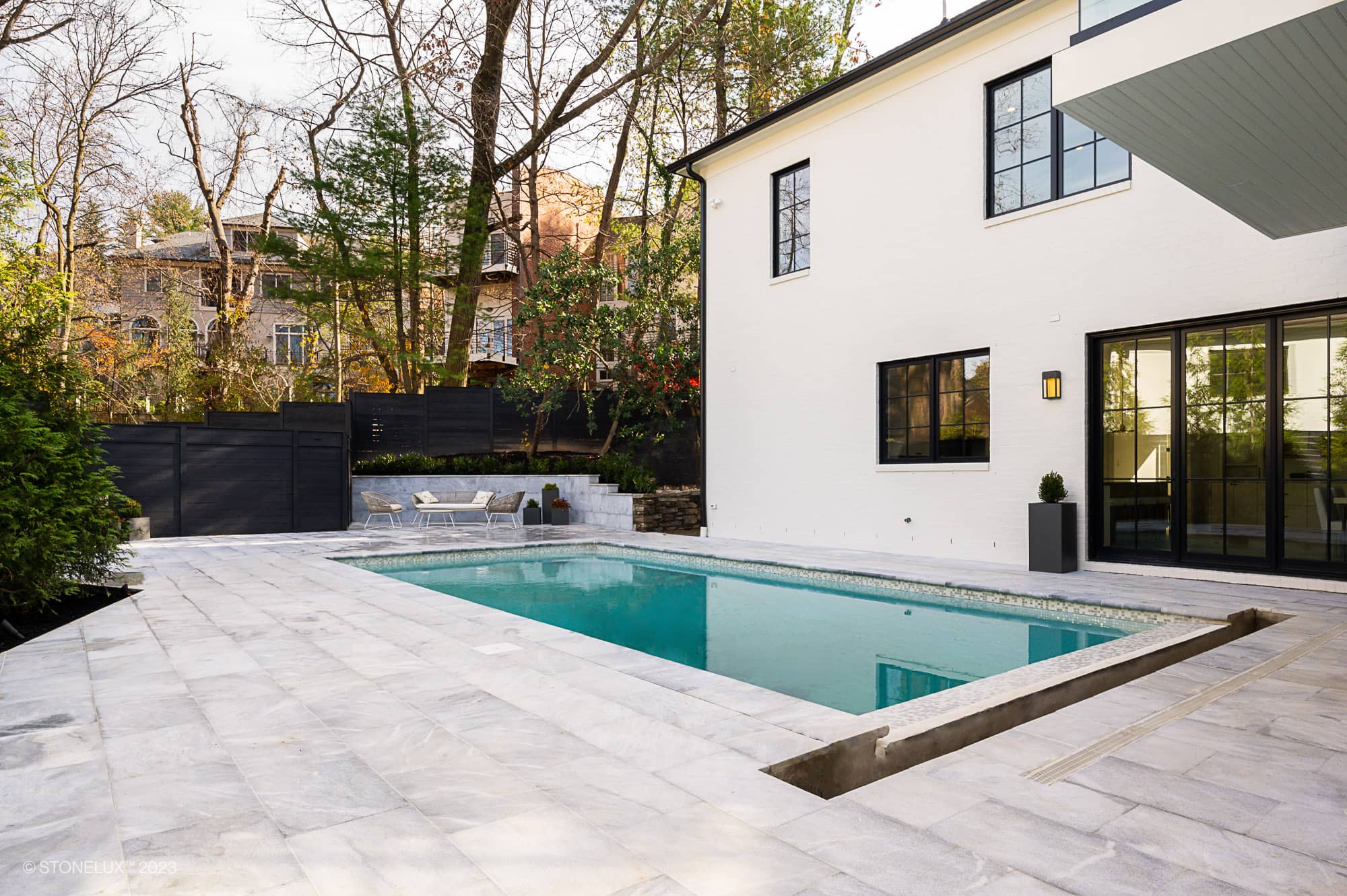 Modern backyard featuring Arctic Elegance: The Blue and Grey Sandblasted Marble Pool and Patio Project, with a rectangular swimming pool, white two-story house, lounge chairs, lush trees, and a sleek black fence.