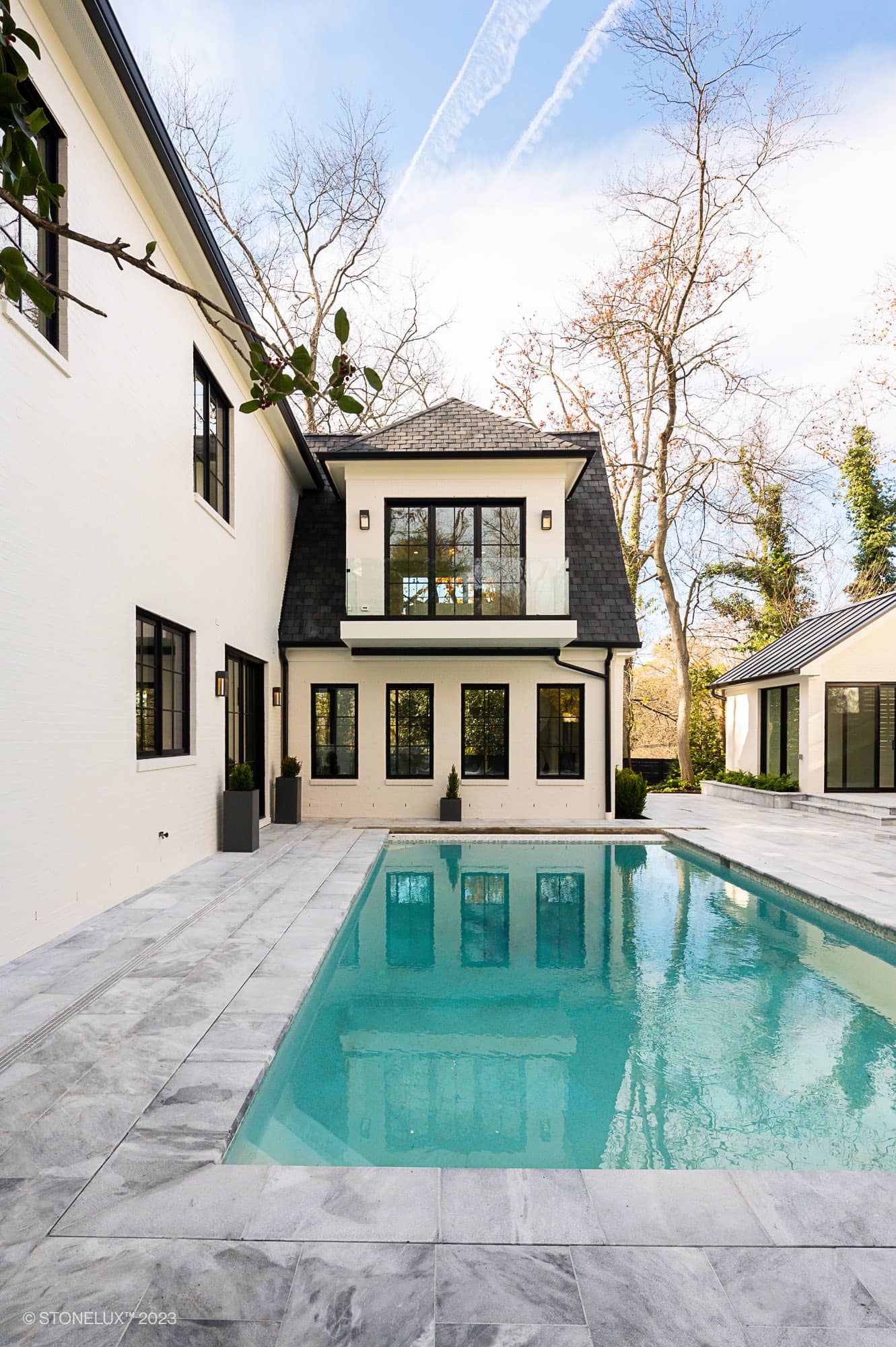 Modern house with cream-colored walls, large windows, and black trim surrounds an Arctic Elegance: The Blue and Grey Sandblasted Marble Pool and Patio Project. Light gray stone tiles border the pool, with leafless trees under a blue sky in the background.