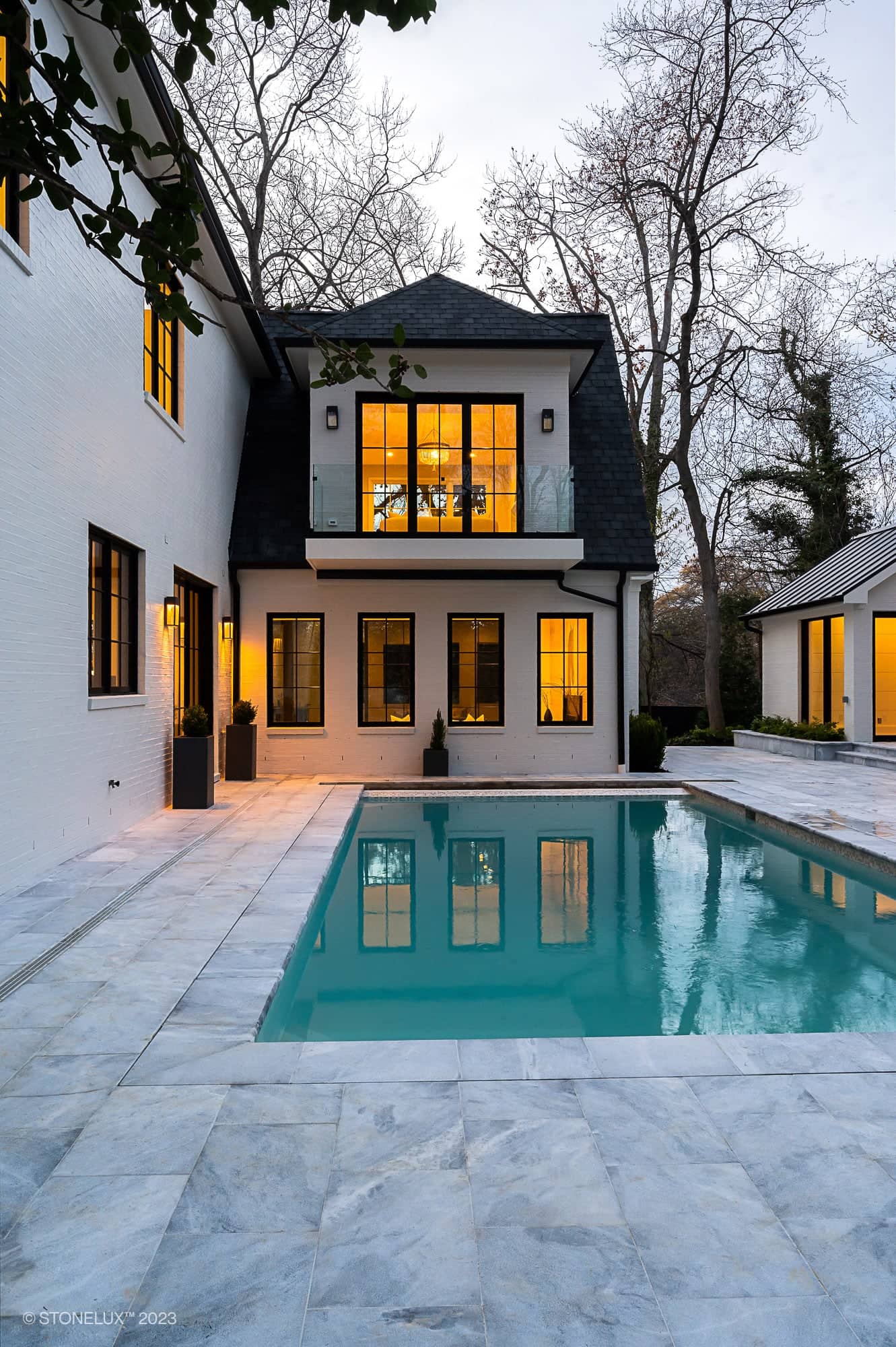 Modern two-story house with white exterior and warm-lit windows, featuring the Arctic Elegance: The Blue and Grey Sandblasted Marble Pool and Patio Project in a spacious stone-tiled backyard, surrounded by leafless trees at dusk.