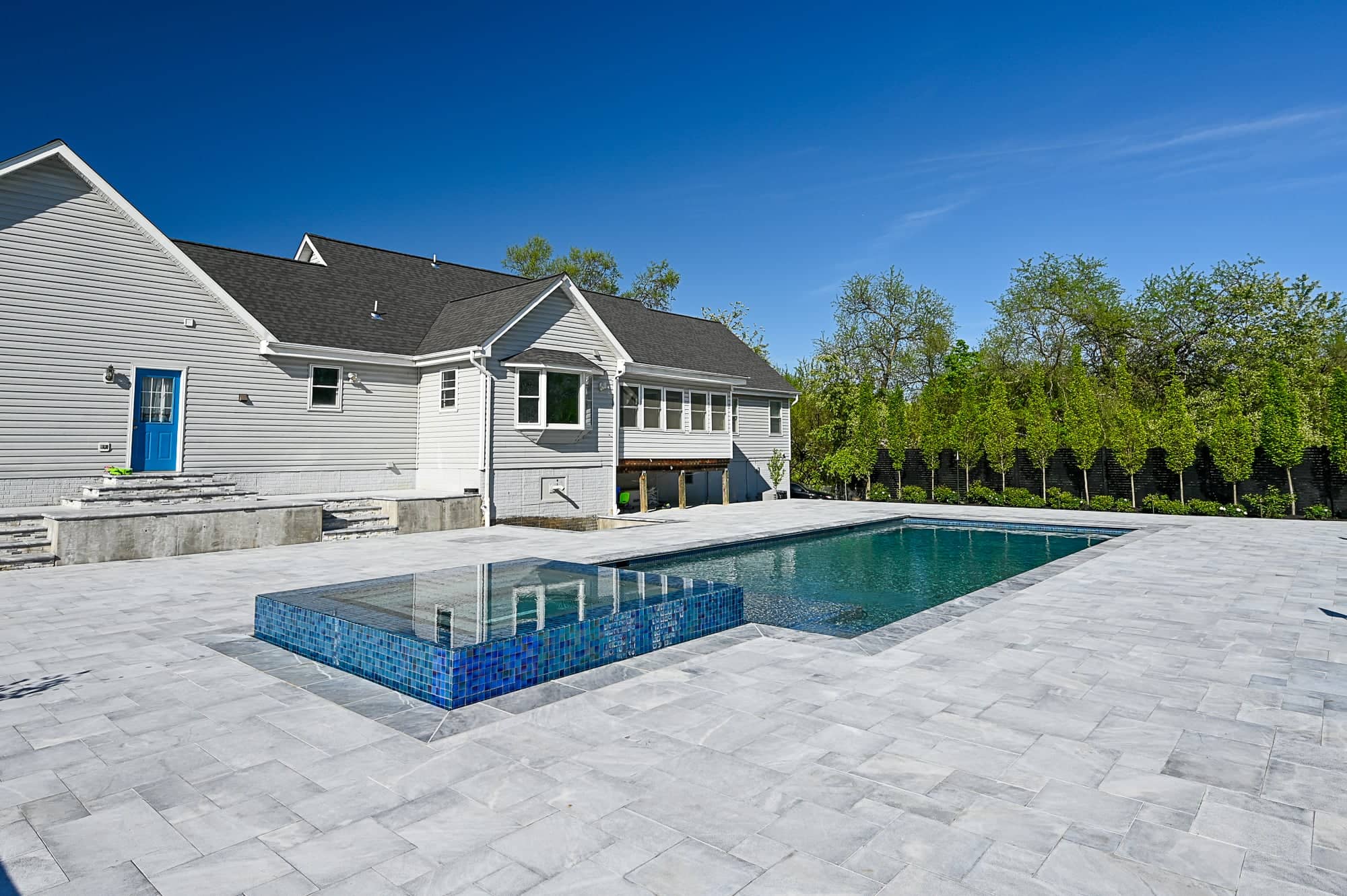 A modern backyard, inspired by Arctic Retreat, features a rectangular swimming pool and raised spa, surrounded by luxury Arctic Grey Marble pavers. The light-colored house with a blue door overlooks trees lining the yard’s edge.