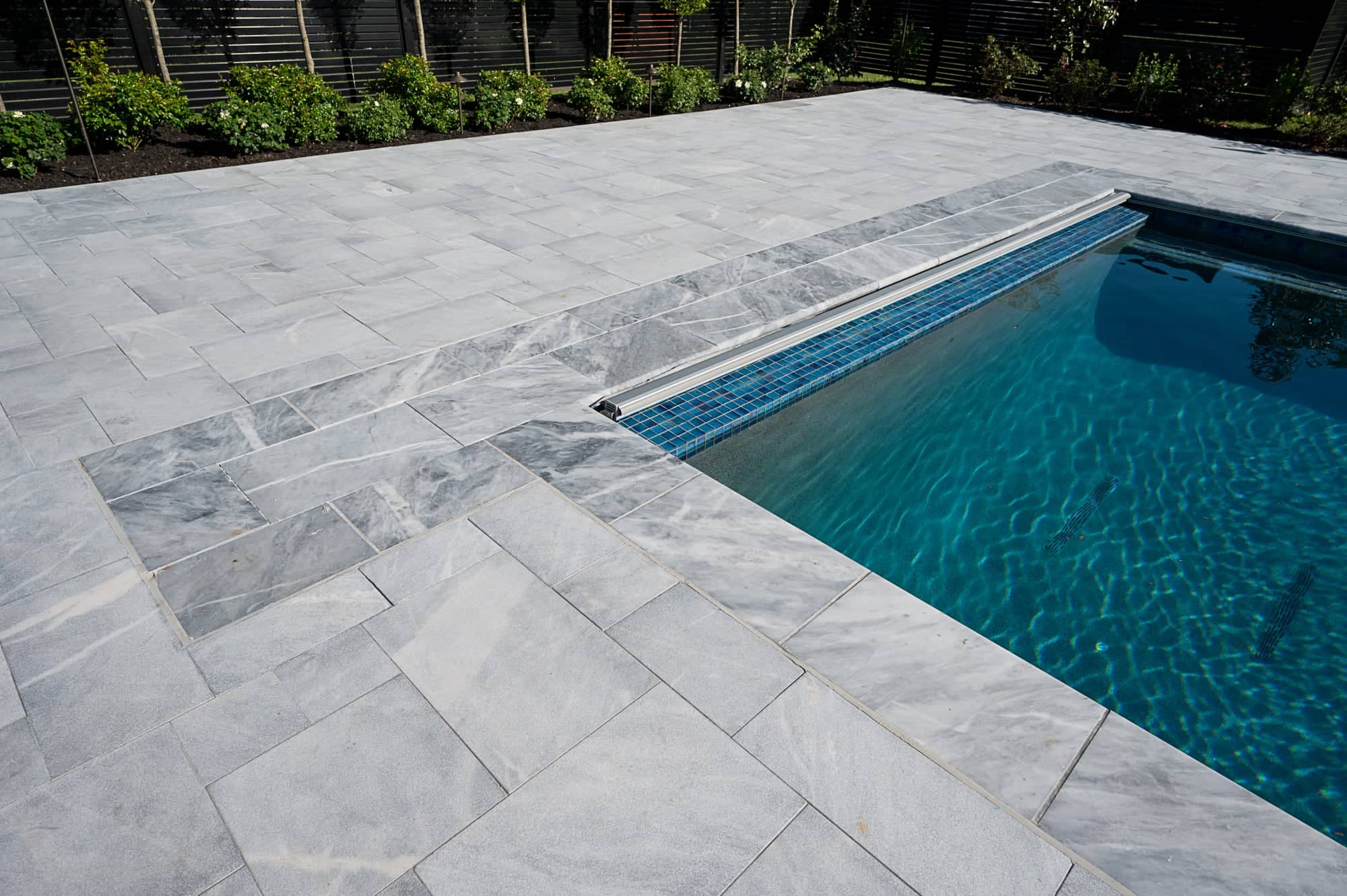 Close-up view of a modern swimming pool with clear blue water, surrounded by Arctic Grey Marble Pavers from the Arctic Retreat: Luxury Pool and Patio collection. Lush green shrubs create a tranquil garden backdrop.