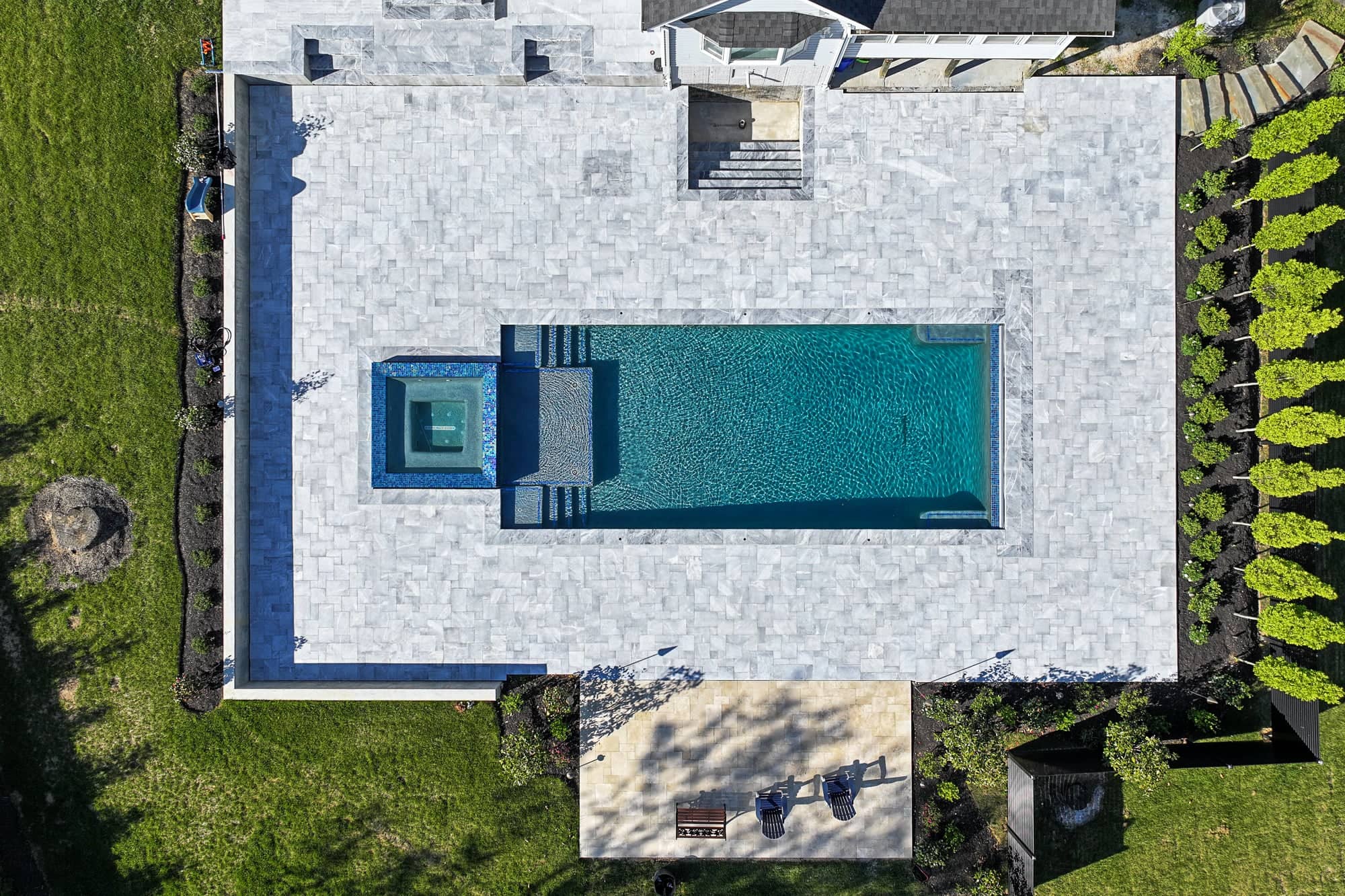 Aerial view of an Arctic Retreat: luxury pool and patio with Arctic Grey Marble Pavers, featuring a rectangular backyard swimming pool, attached square hot tub, lush grass, pristine landscaping, two lounge chairs, and a small table nearby.