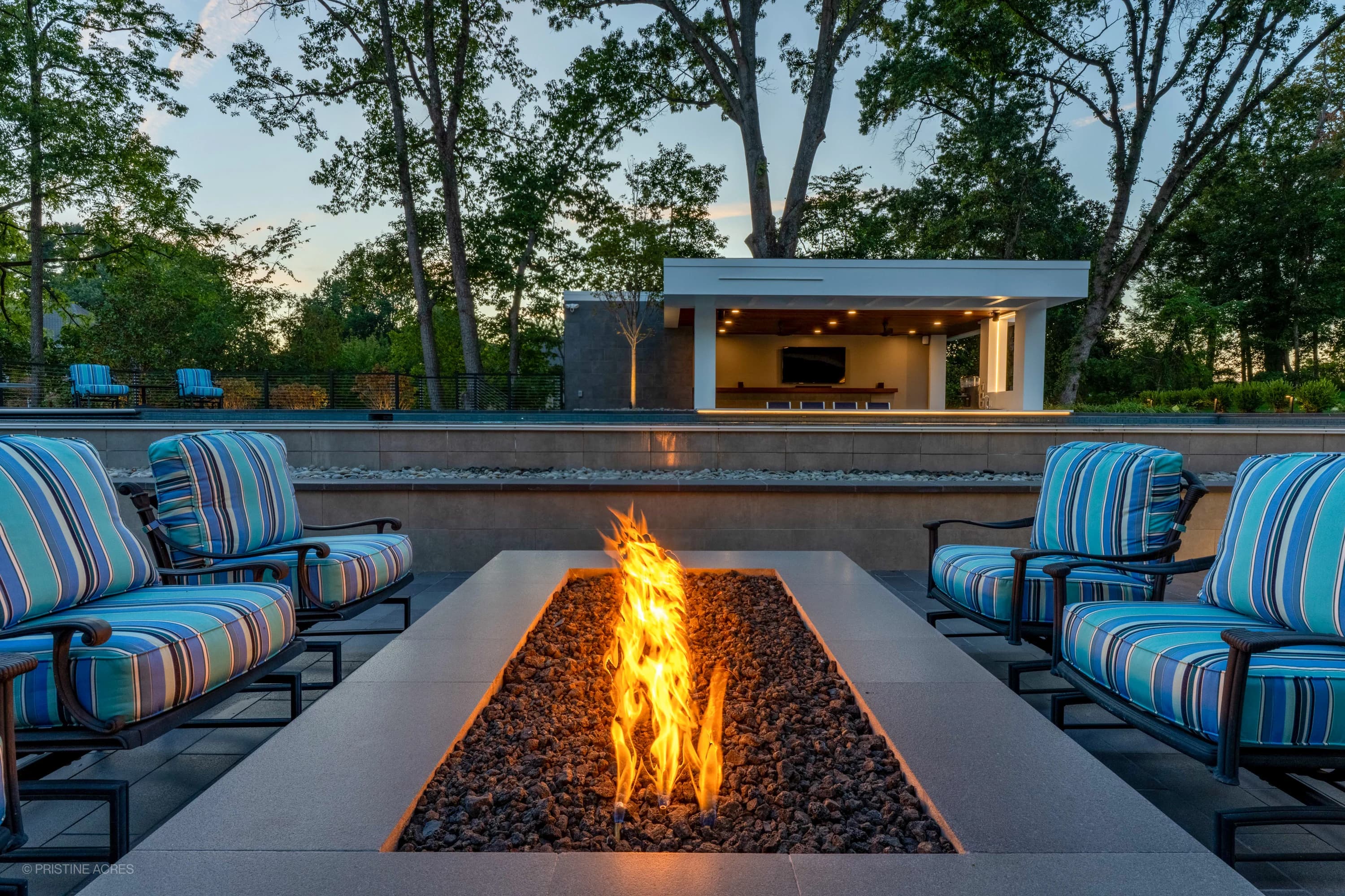 Modern McLean pool project featuring Black Basalt and Nexus White Porcelain collections. A sleek fire pit with blue-striped chairs enhances the contemporary design. Surrounded by lush greenery and a stylish pool house, creating a serene backyard atmosp...