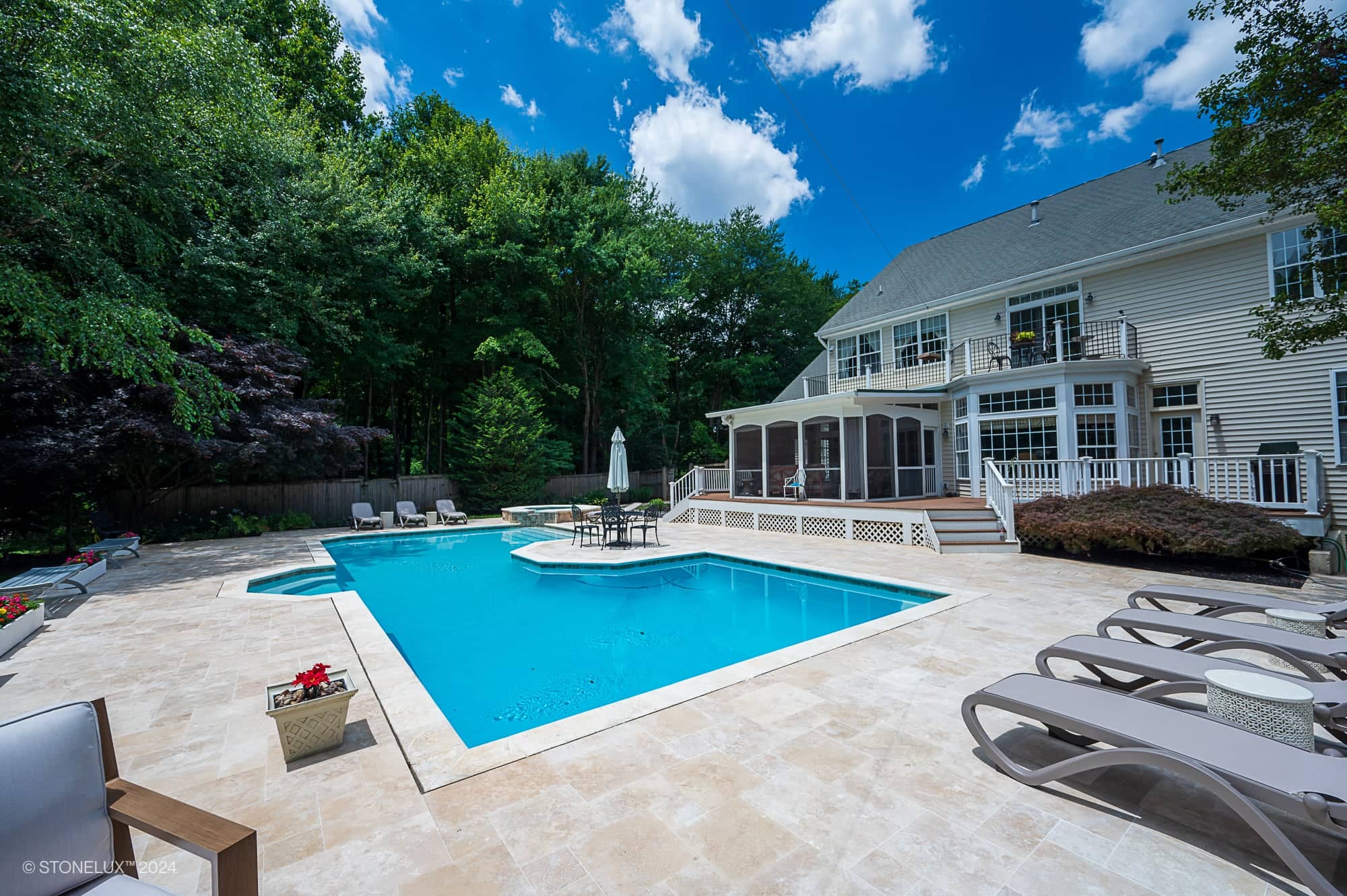 A spacious backyard features an Ivory Travertine retreat pool under the trees, with a small island, lounge chairs, potted plants, and a two-story white house with a screened porch set against a blue sky.