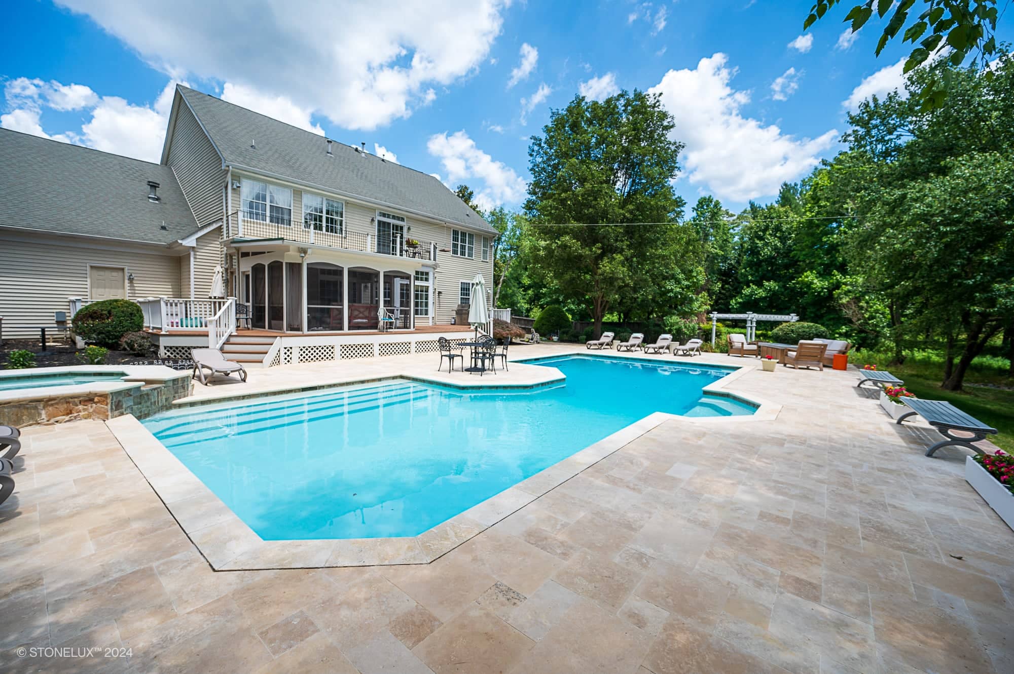 Spacious backyard with a large, light blue swimming pool surrounded by an ivory travertine stone-tiled deck. Lounge chairs and patio furniture create a retreat pool under the trees, with a two-story house in the background.