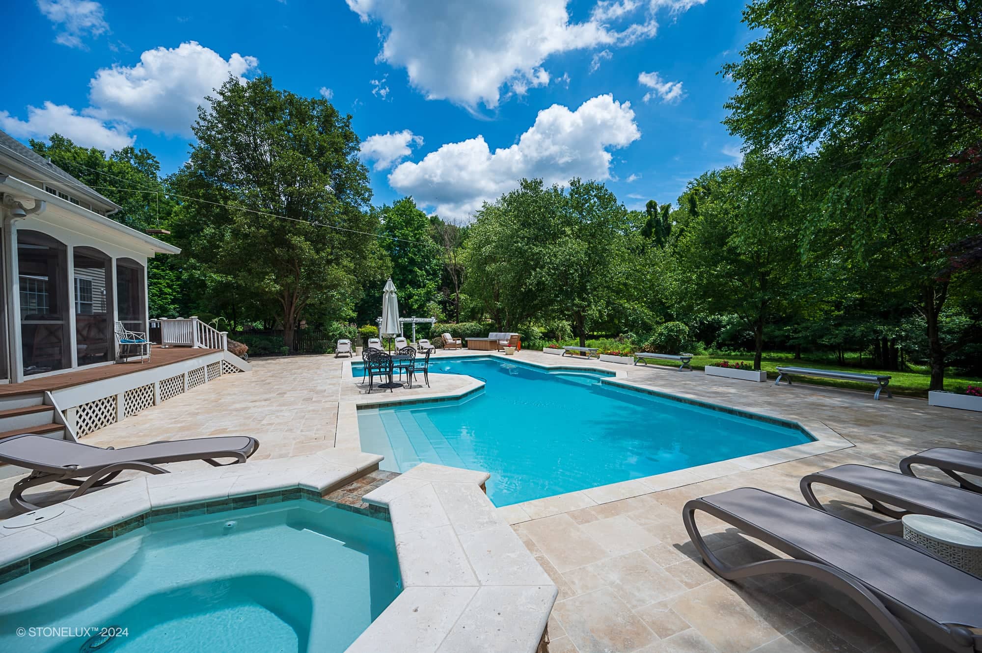 A luxurious backyard featuring an Ivory Travertine retreat pool under the trees, complete with a hot tub, sun loungers, outdoor dining table, all beneath a vibrant blue sky with fluffy clouds.