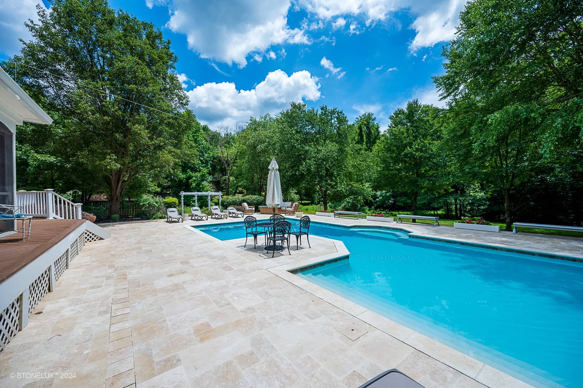 A spacious backyard features a large swimming pool with ivory travertine decking—a true retreat pool under the trees—surrounded by lounge chairs, lush greenery, and a dining table with an umbrella beneath a bright blue sky.
