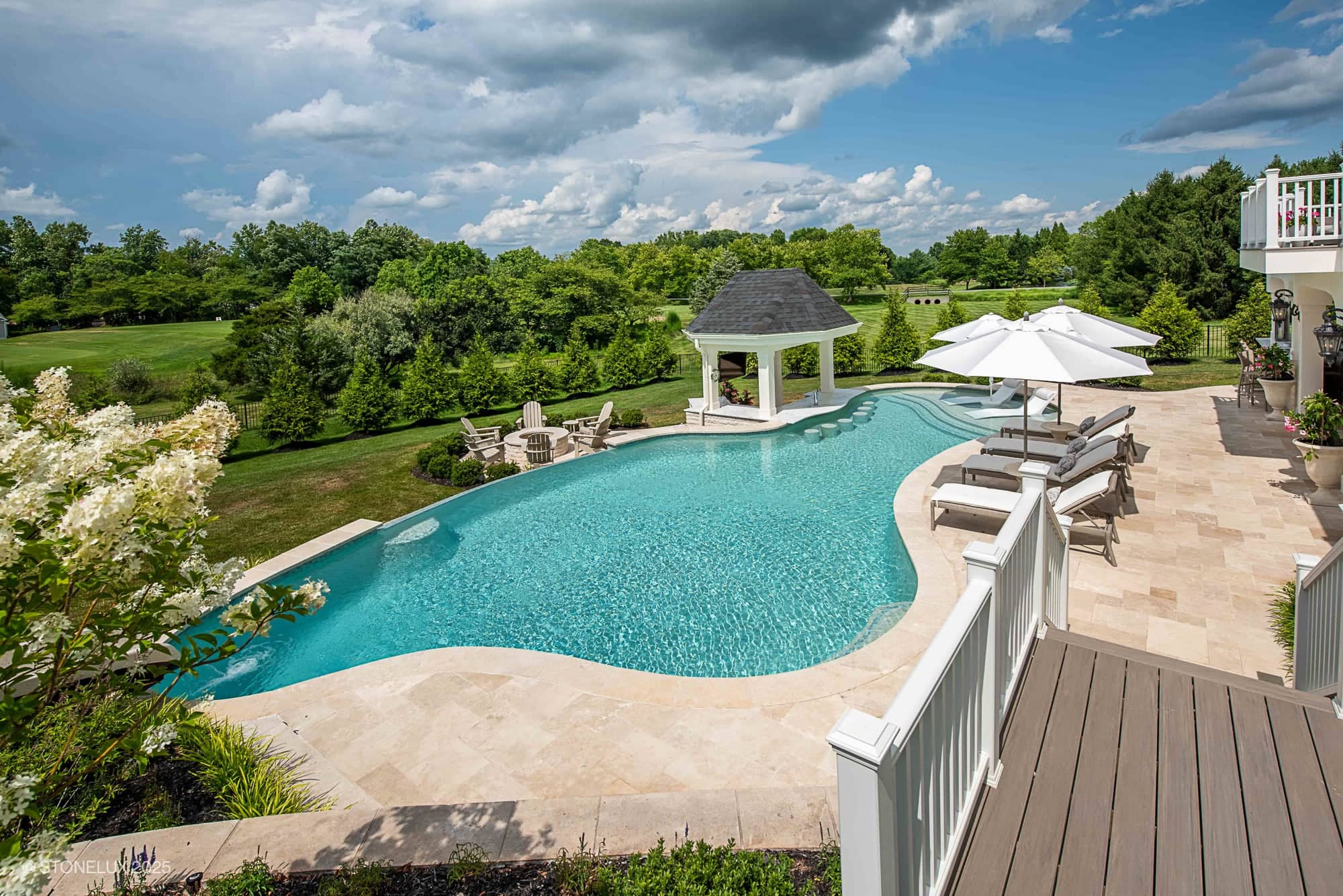 Backyard with a large, curving Leesburg Grecian Oasis – Ivory Travertine Pool Project surrounded by lounge chairs and umbrellas, a poolside gazebo, stone patio, green lawn, and trees under a partly cloudy sky.