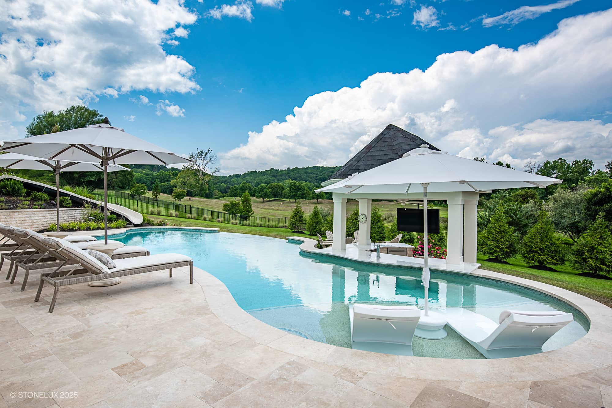 Experience the Leesburg Grecian Oasis – an ivory travertine pool project featuring lounge chairs, umbrellas, a waterslide, and a covered outdoor TV area, all nestled in lush greenery with open field views under a partly cloudy sky.