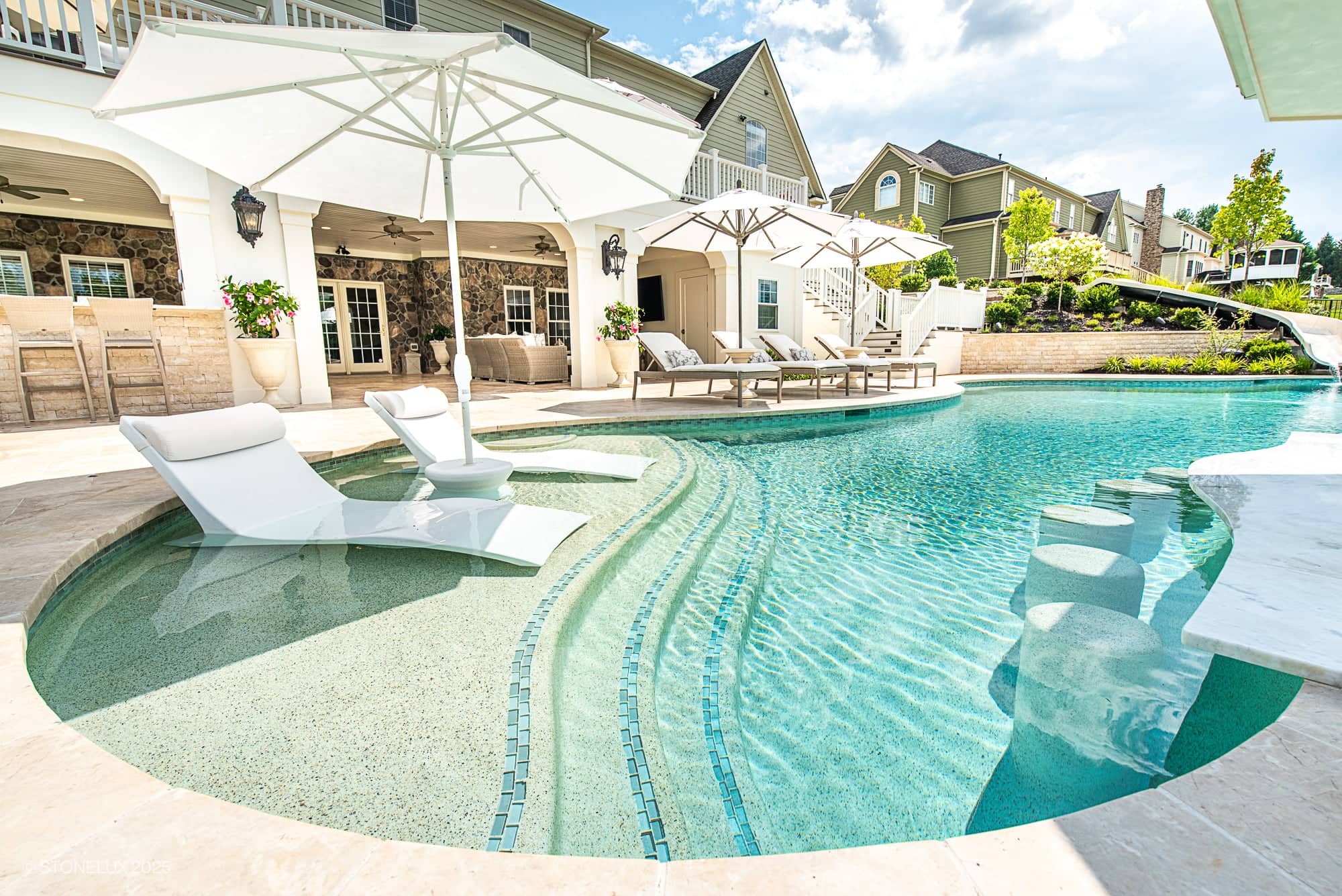 Experience the Leesburg Grecian Oasis – Ivory Travertine Pool Project: a luxury backyard retreat with lounge chairs in the shallow end, elegant white umbrellas, a swim-up bar, waterslide, and modern patio furniture—all under a brilliant blue sky.