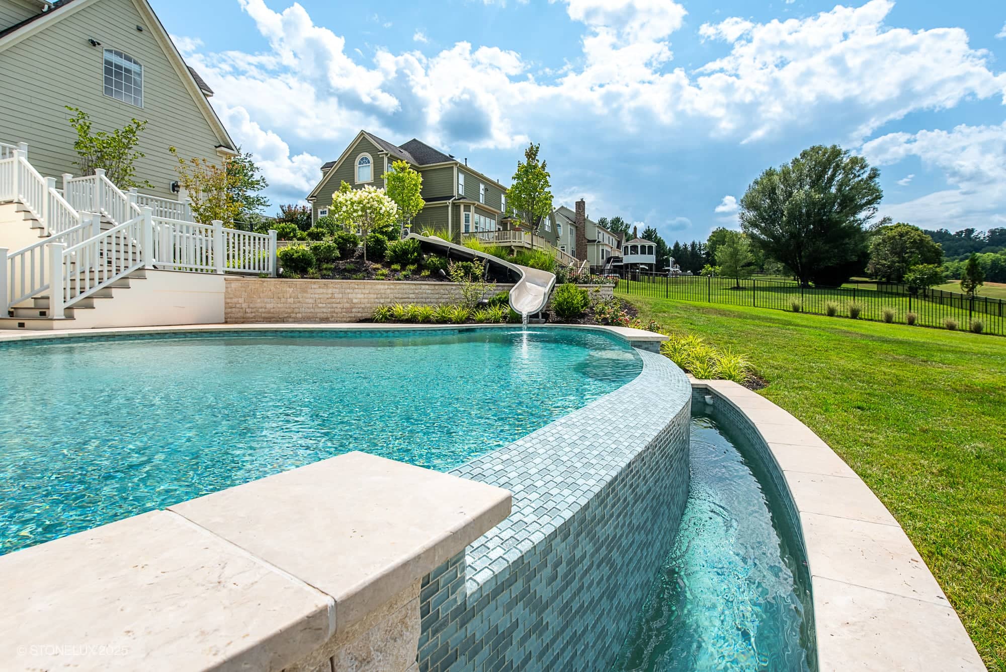 A backyard features the Leesburg Grecian Oasis – Ivory Travertine Pool Project, showcasing an infinity-edge swimming pool with clear water, a curved stone tile border, a waterslide, lush greenery, and a large grassy lawn by suburban homes.