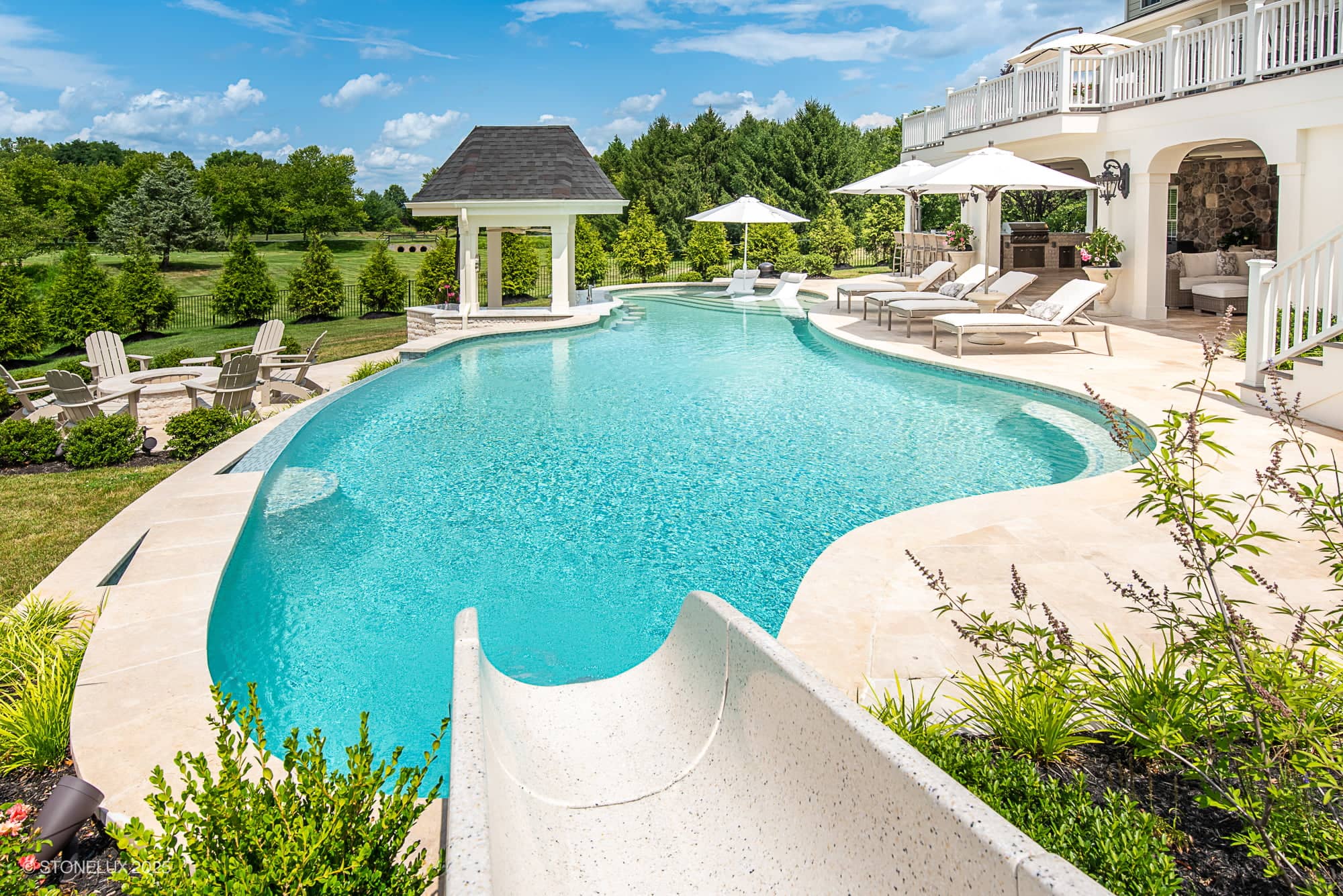 Experience the Leesburg Grecian Oasis – Ivory Travertine Pool Project: a luxurious backyard pool with a water slide, lounge chairs, umbrellas, covered seating area, and lush landscaping set against trees and a spacious lawn on a sunny day.