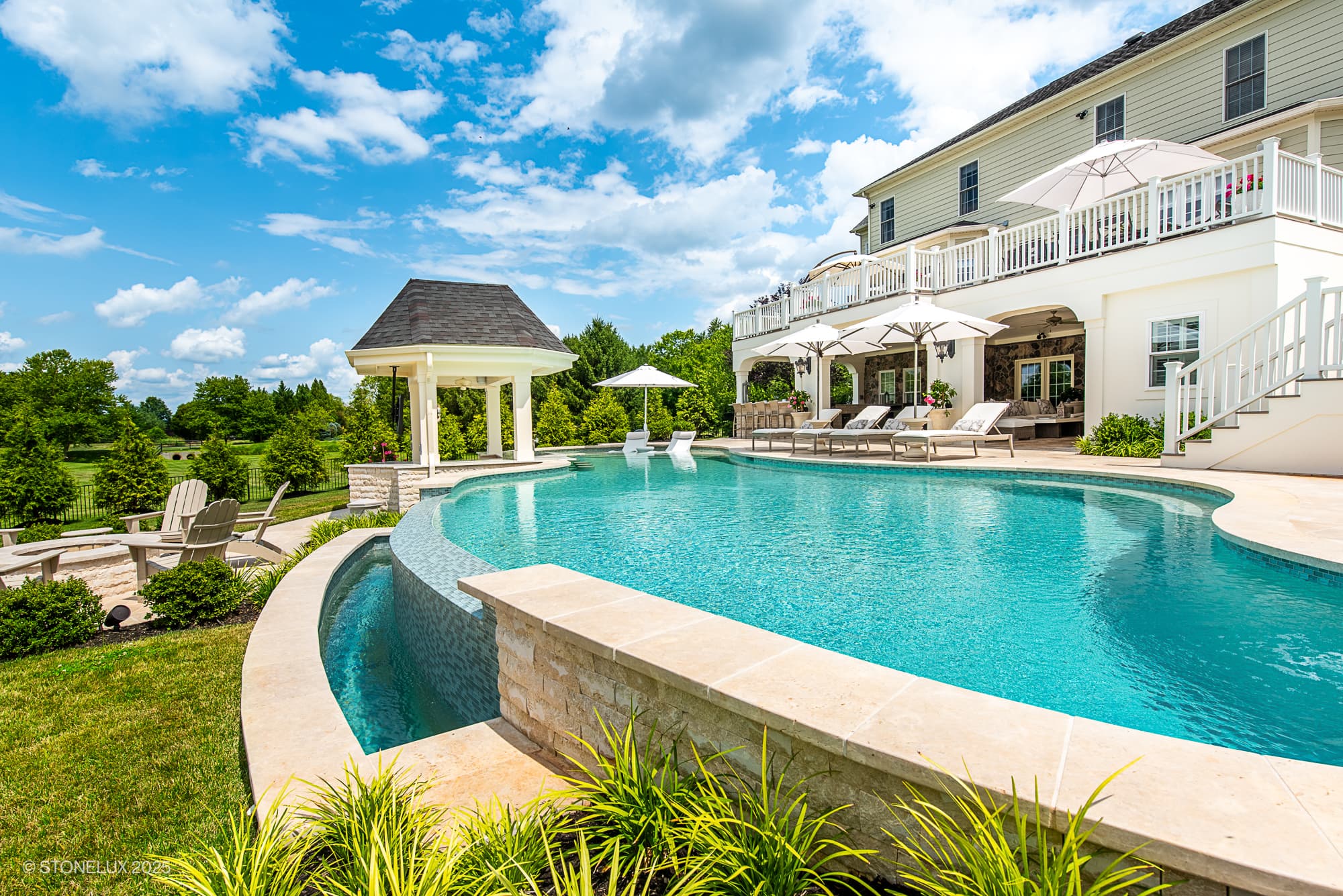 Luxury backyard featuring the Leesburg Grecian Oasis – Ivory Travertine Pool Project, with a large curved swimming pool, shaded lounge chairs, gazebo, white two-story house, green lawn, trees, and Adirondack seating under blue skies.