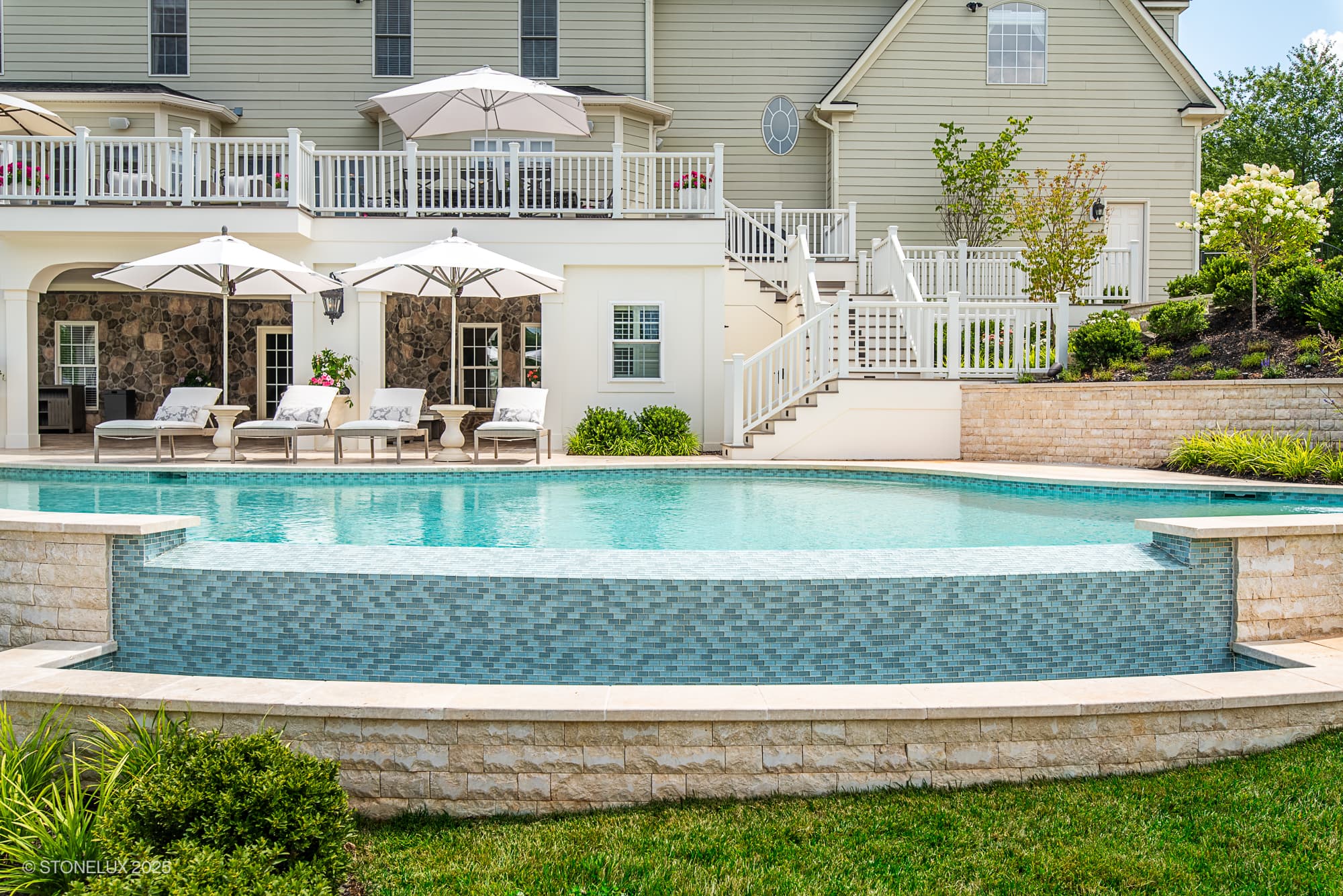 A luxurious backyard, part of the Leesburg Grecian Oasis – Ivory Travertine Pool Project, features an infinity pool with mosaic tiles, lounge chairs under white umbrellas, a patio area, and a large beige house with balconies and white railings.