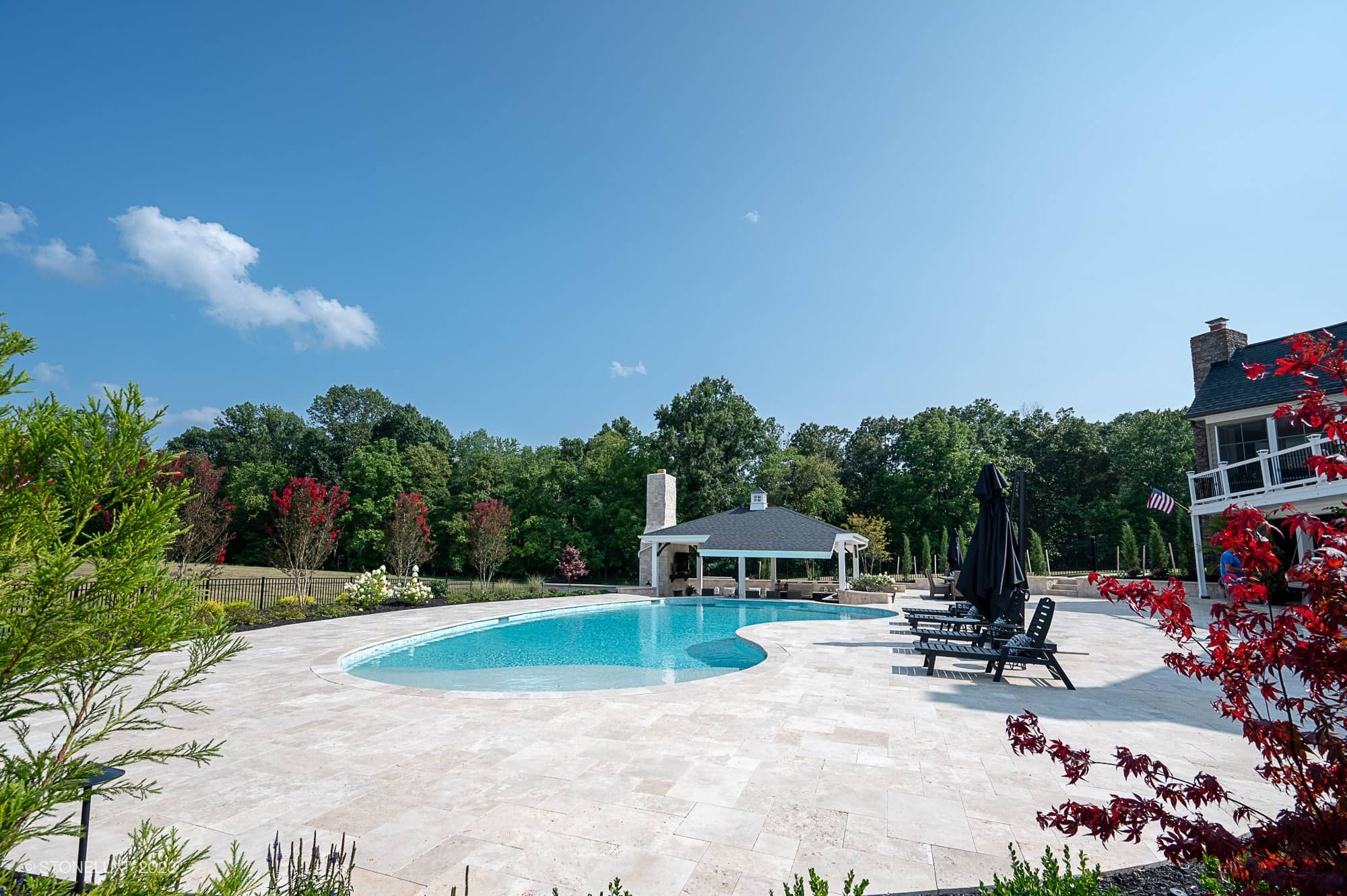 A spacious backyard featuring a circular swimming pool with Beige Bliss: Luxurious Travertine Pool and Pool House, lounge chairs, a covered patio with a fireplace, and lush greenery under a clear blue sky.