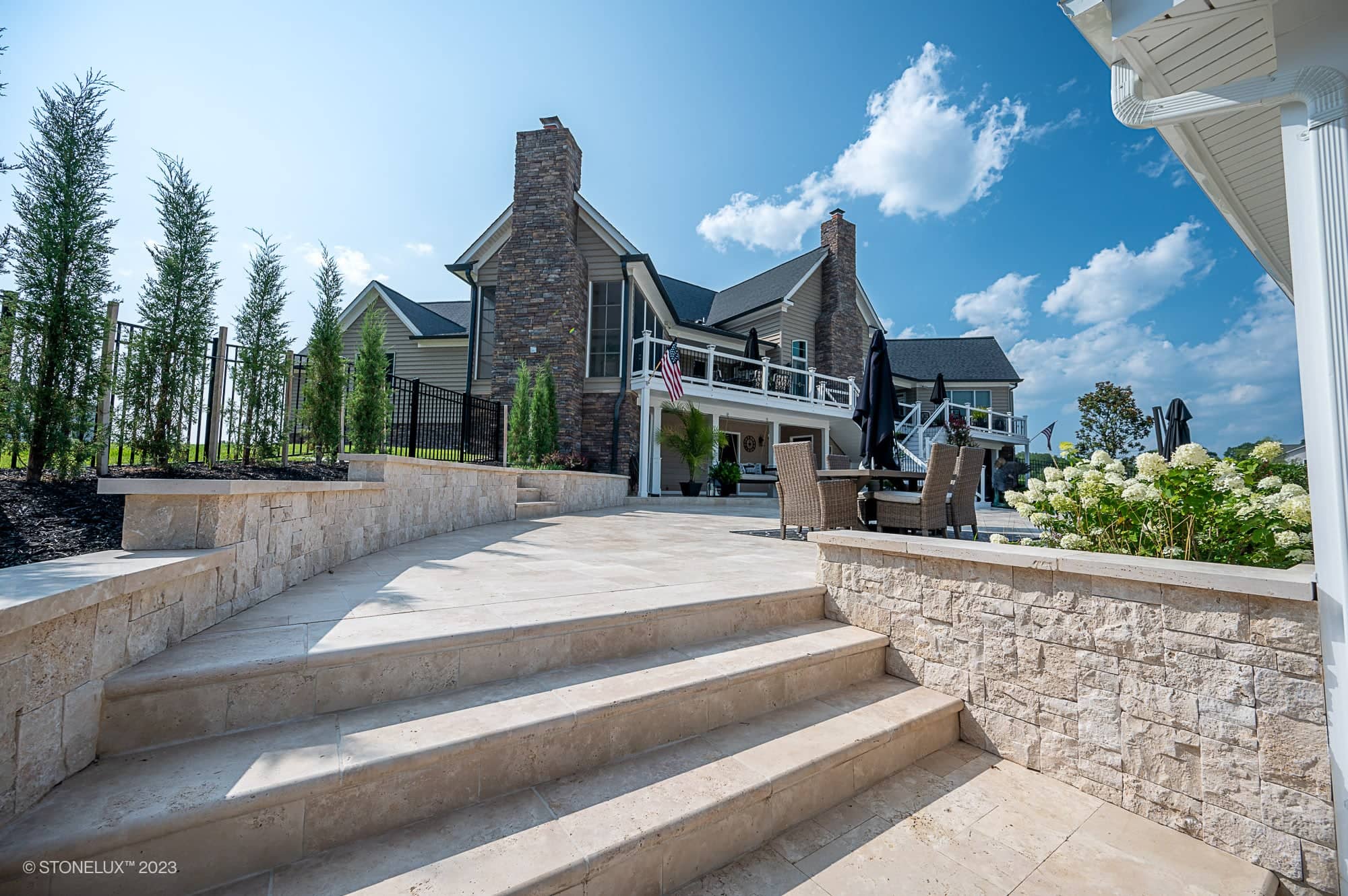 Elegant stone steps lead to a spacious backyard patio with outdoor dining furniture, surrounded by landscaping and tall greenery—a true Beige Bliss: Luxurious Travertine Pool and Pool House complete the scene with a blue sky in the background.