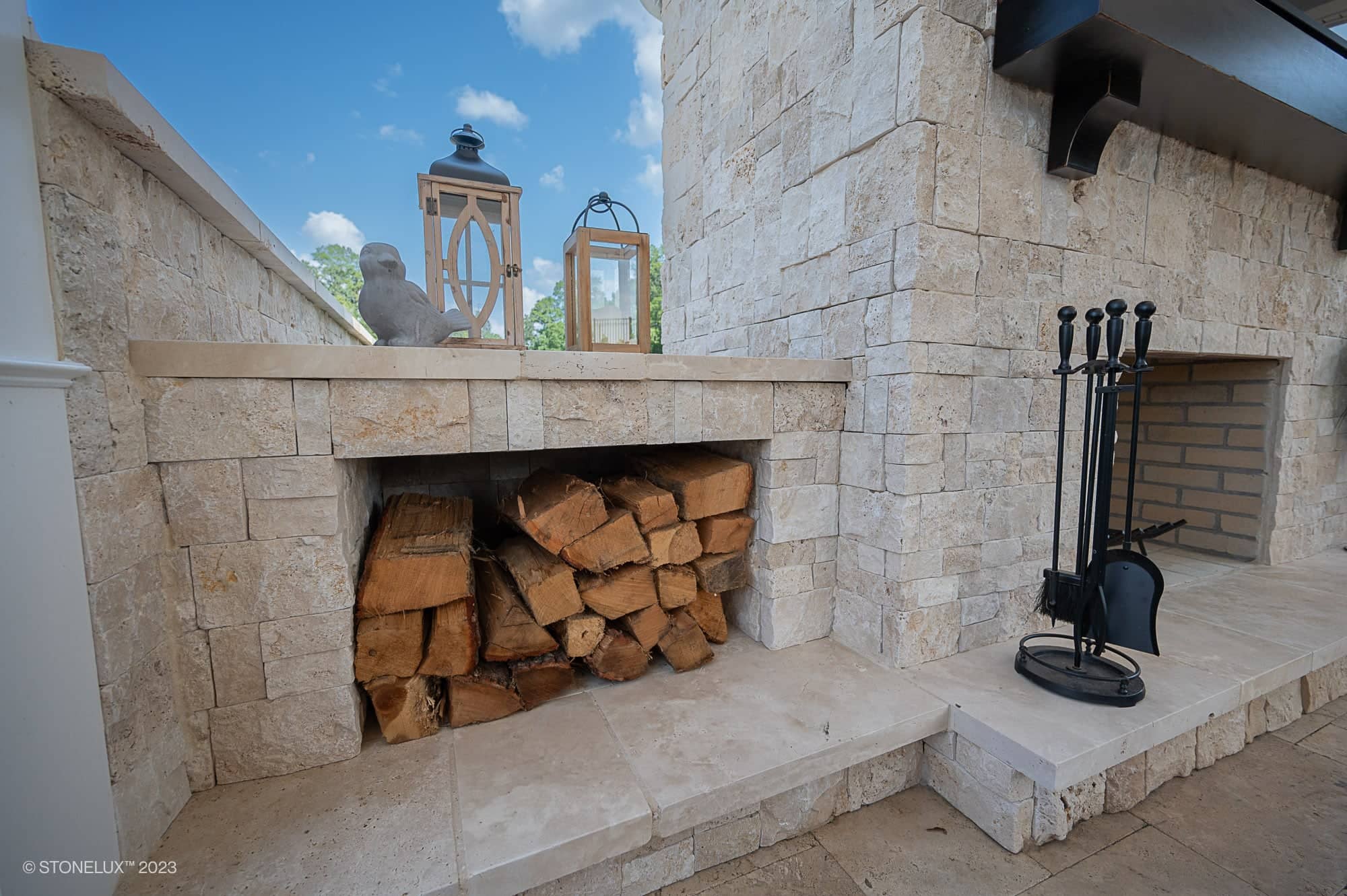 A stone outdoor fireplace with a storage area filled with stacked firewood, two lanterns, and a decorative bird figure on the mantel creates Beige Bliss beside the luxurious travertine pool and pool house. Blue sky and trees complete the serene setting.