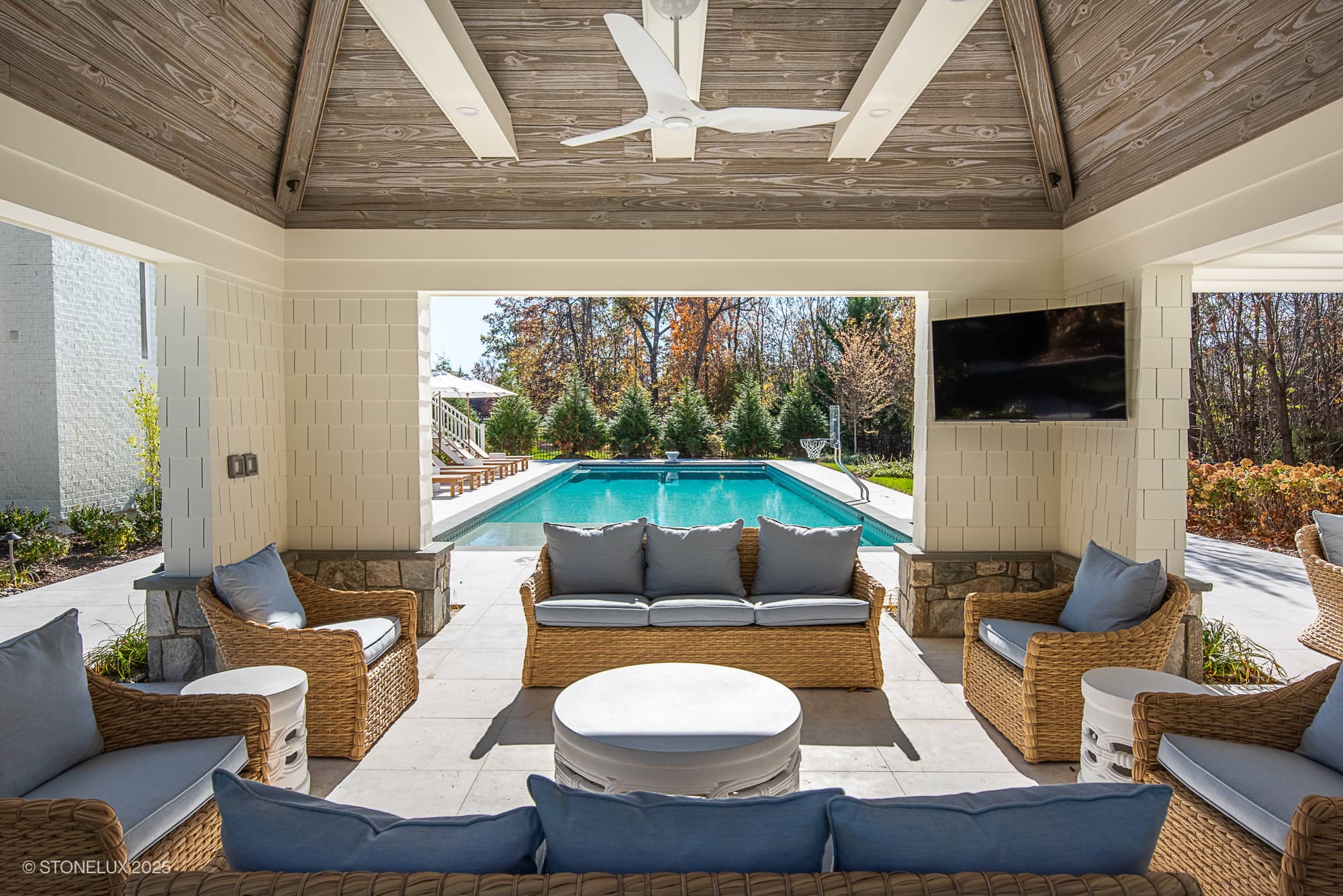 Covered outdoor seating area with wicker furniture and blue cushions, facing a swimming pool. Enjoy Ashburn Family Fun and elegant outdoor living in Pera Cream Marble, with ceiling fan, TV, and views of trees and patio in the background.