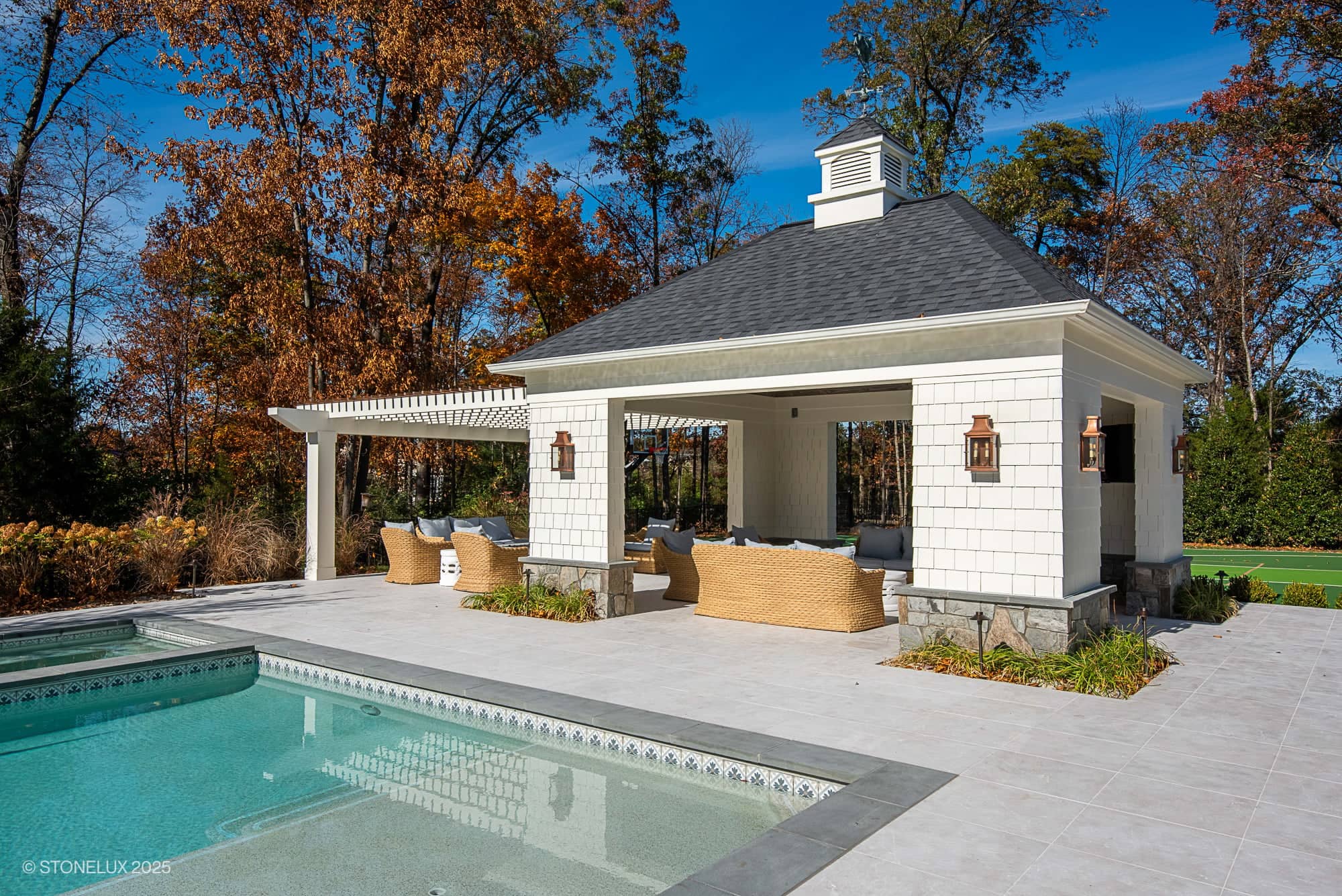 A modern poolside pavilion with a shingled roof and white exterior sits near a swimming pool, surrounded by wicker furniture and autumn trees—perfect for Ashburn Family Fun and elegant outdoor living in Pera Cream Marble under a bright blue sky.