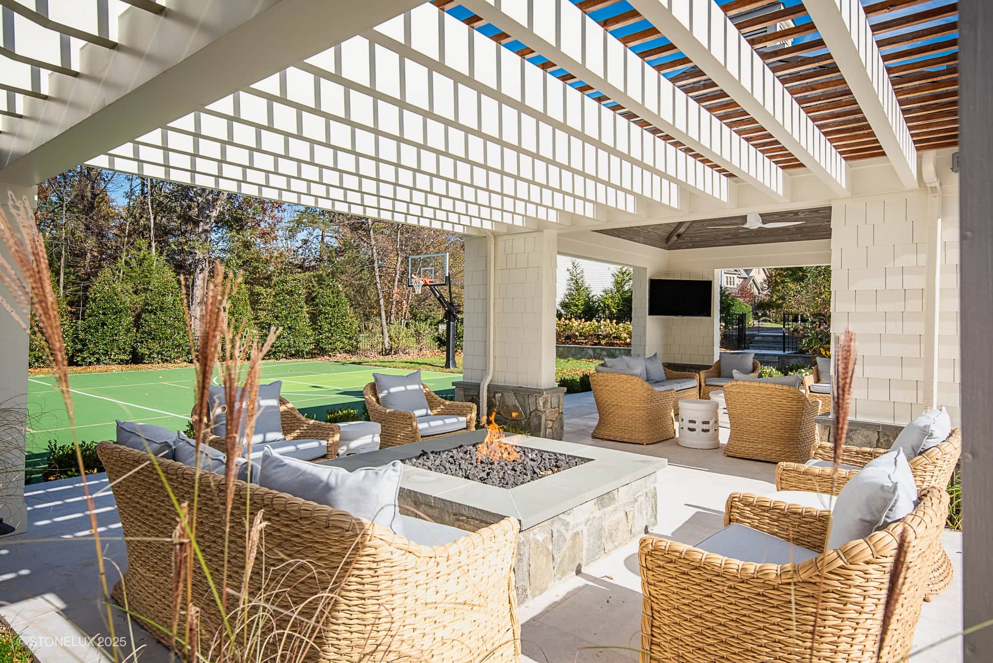 Experience Ashburn Family Fun in this elegant outdoor living space, featuring wicker chairs on Pera Cream Marble under a white pergola. Enjoy the stone firepit, TV, and basketball court—all surrounded by tall trees.