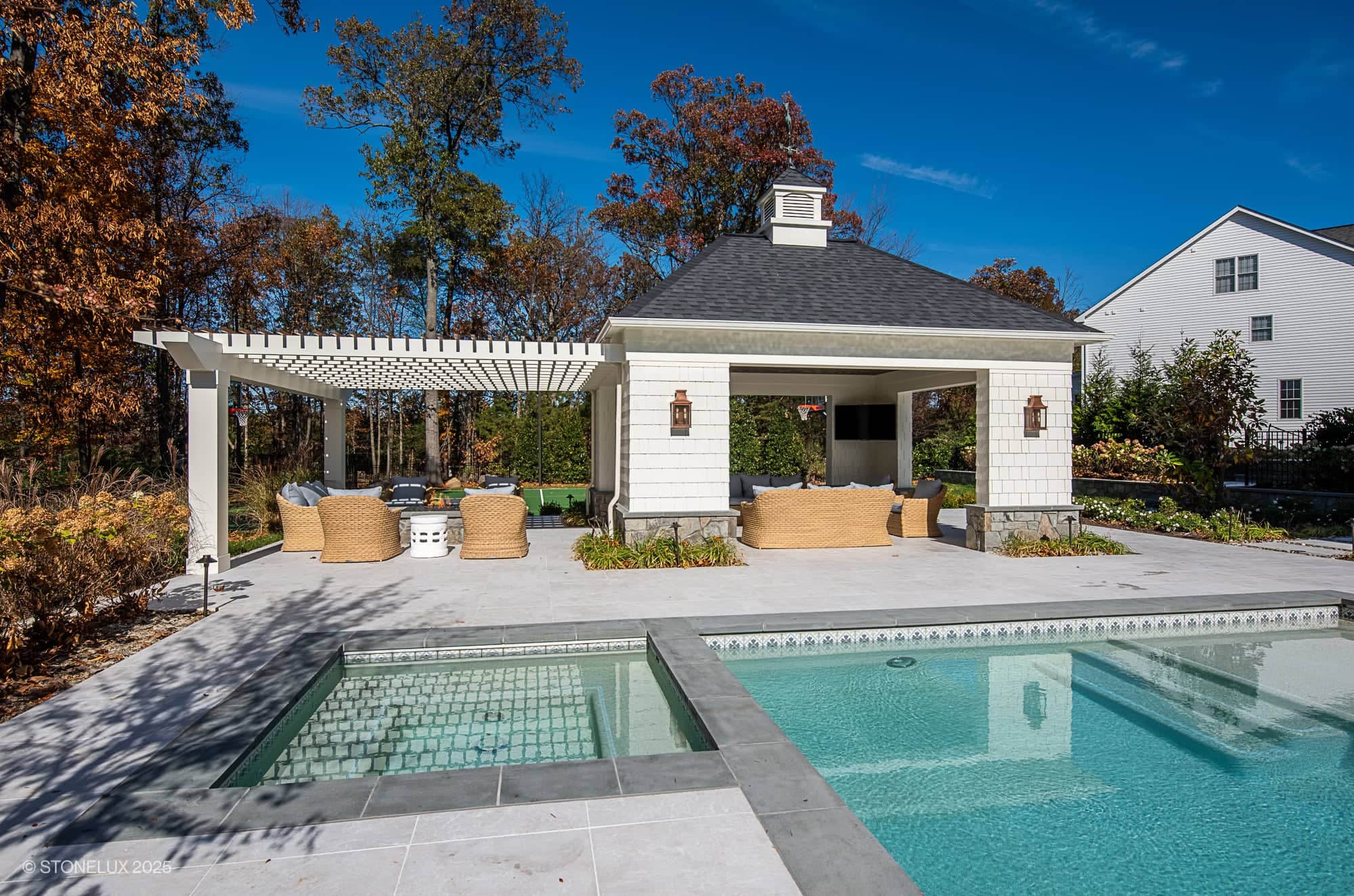 Outdoor pool area perfect for Ashburn Family Fun, featuring elegant outdoor living in Pera Cream Marble. Enjoy the cabana with wicker seating, a white pergola, hot tub, and lush landscaping under a clear blue sky.