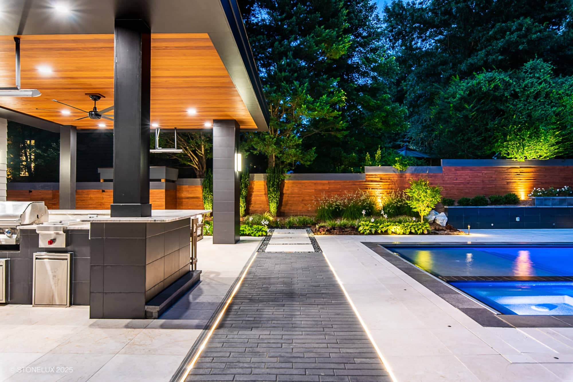 Modern outdoor kitchen and lounge area beside a lit swimming pool, featuring McLean Modern Zen – Outdoor Living in Pera Cream Marble & Black Basalt, with sleek countertops, a barbecue grill, wood ceiling accents, landscaped greenery.