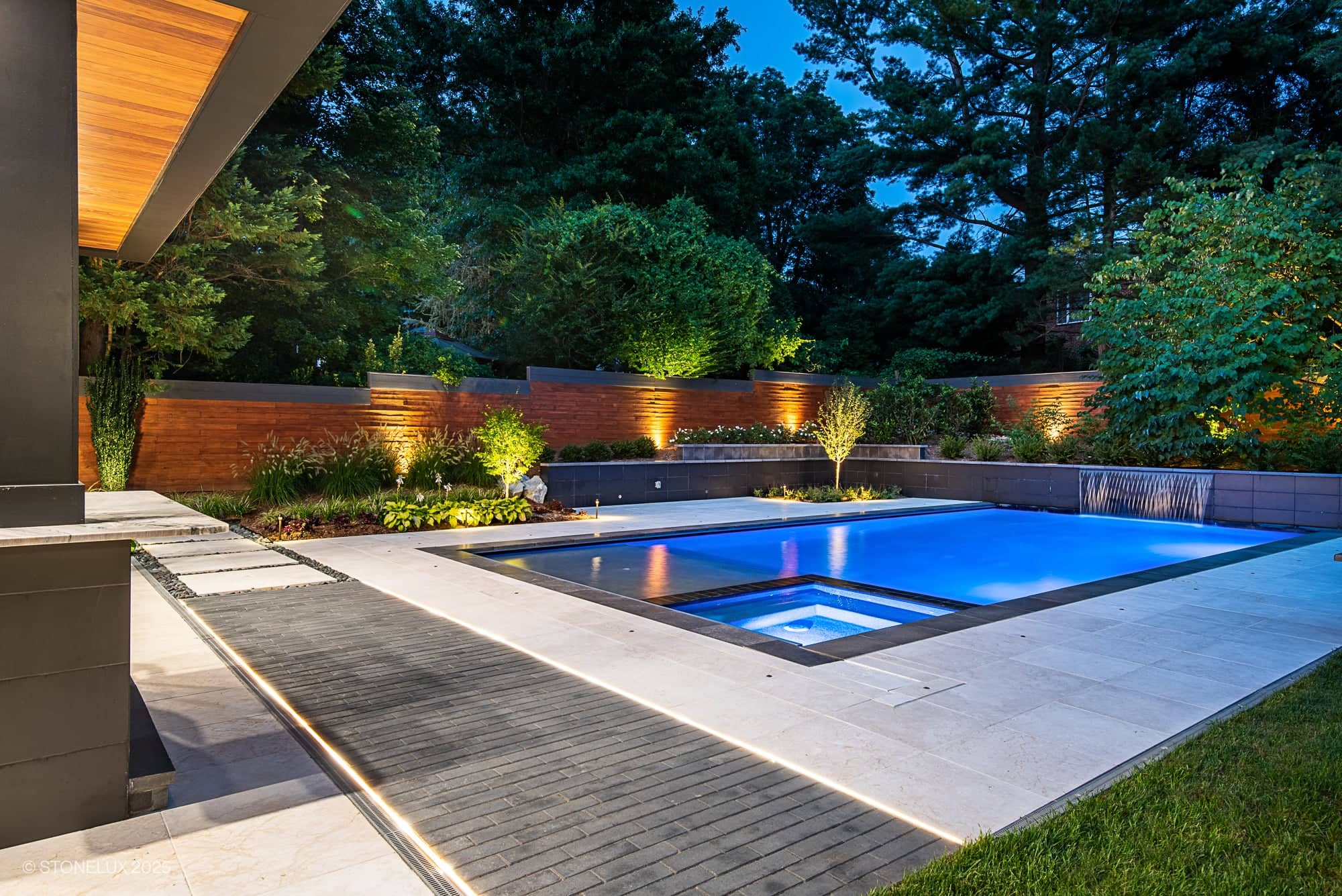 Modern backyard with a lit swimming pool and hot tub at dusk, featuring McLean Modern Zen – Outdoor Living in Pera Cream Marble & Black Basalt, surrounded by trees, fencing, landscaped plants, and stone patio tiles for a tranquil retreat.