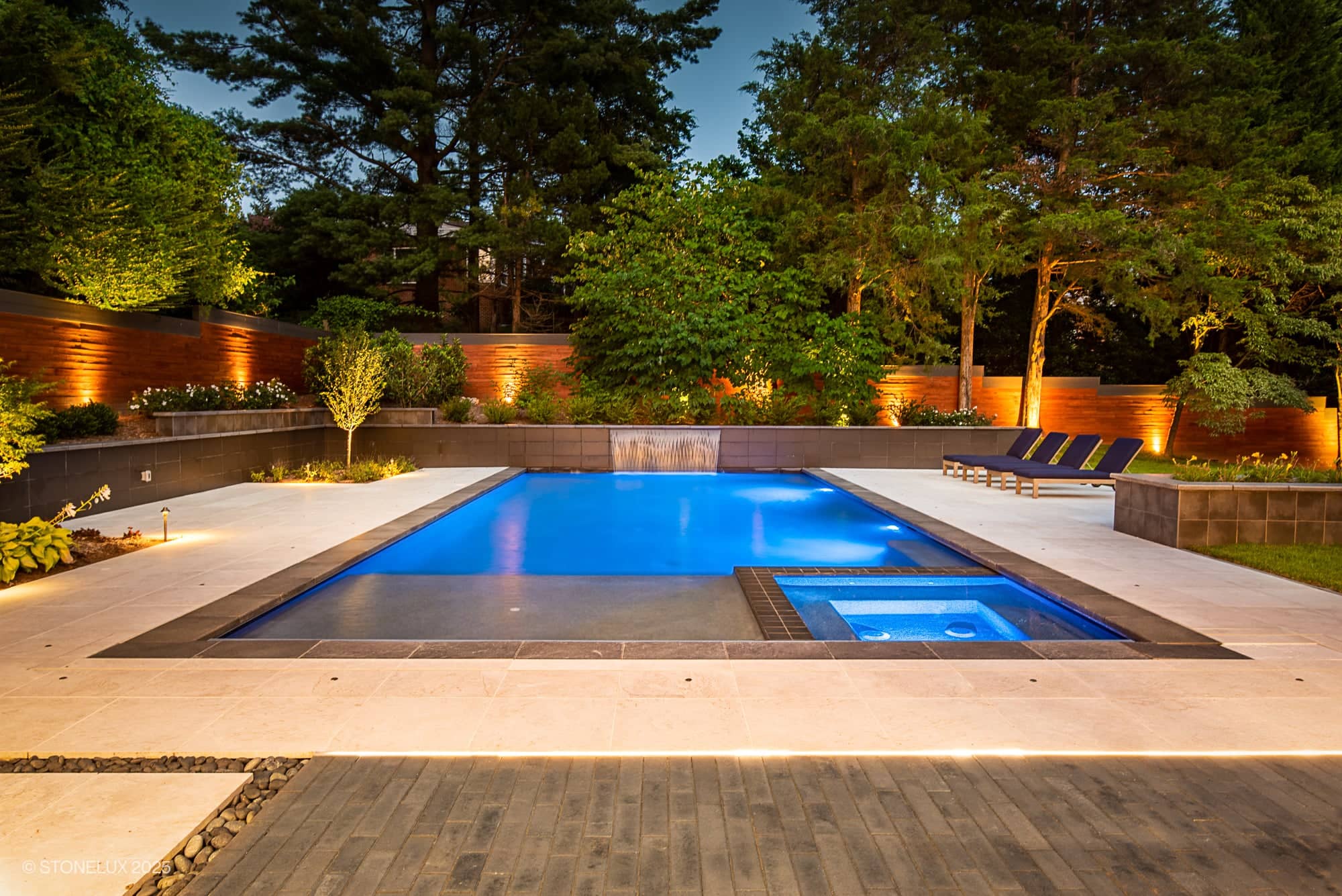 A modern backyard pool at dusk, surrounded by trees and greenery, features McLean Modern Zen – Outdoor Living in Pera Cream Marble & Black Basalt, with underwater lighting, a hot tub, lounge chairs, and a wooden privacy fence with warm accent lights.