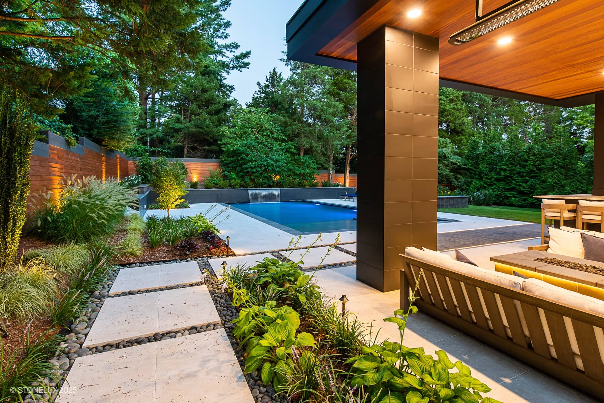 A modern backyard with a covered patio overlooking a rectangular pool, featuring McLean Modern Zen – Outdoor Living in Pera Cream Marble & Black Basalt. Warm outdoor lighting highlights manicured plants and tall trees, creating a cozy atmosphere at dusk.