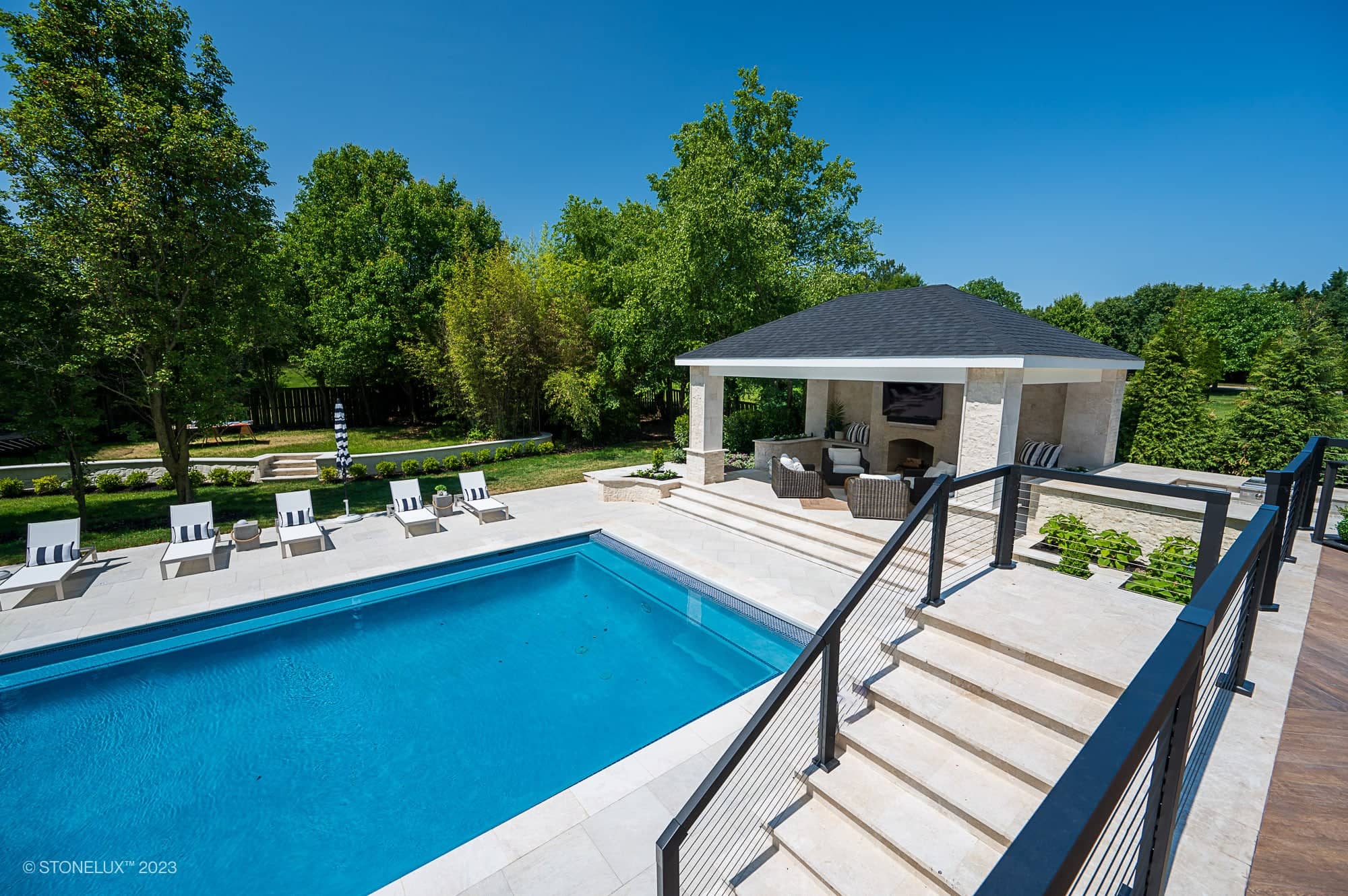 Luxurious backyard with a large rectangular swimming pool featuring White Harmony: Pera Cream Sandblasted Marble, several lounge chairs, a covered outdoor seating area, stone patio, and lush green trees under a clear blue sky.