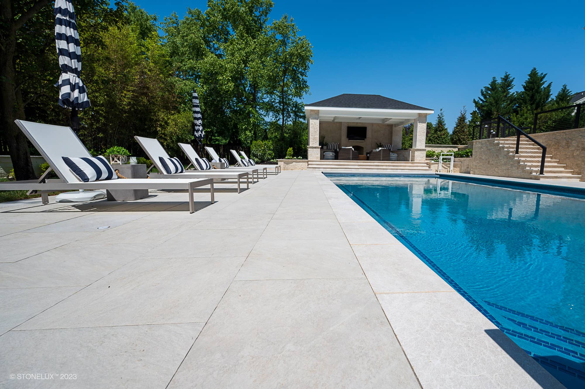 Modern outdoor pool area with White Harmony: Pera Cream Sandblasted Marble tiles, white lounge chairs, striped cushions, and umbrellas. The porcelain poolside retreat is enhanced by a stylish pool house, lush greenery, and clear blue skies.