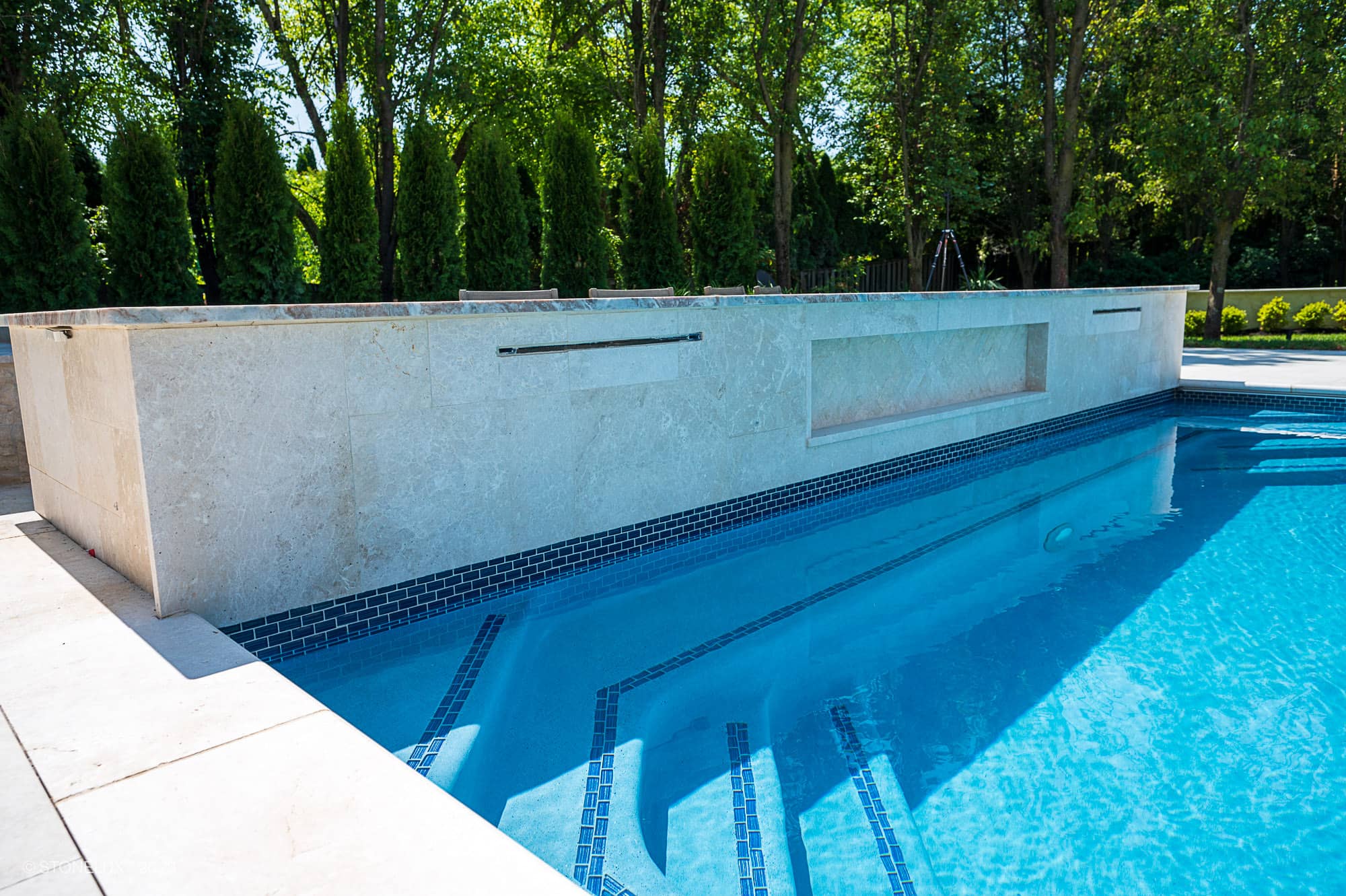 A modern outdoor swimming pool with blue water, mosaic tile accents, and built-in steps features a White Harmony: Pera Cream Sandblasted Marble stone wall beside the pool, surrounded by green trees and landscaping for a serene retreat.