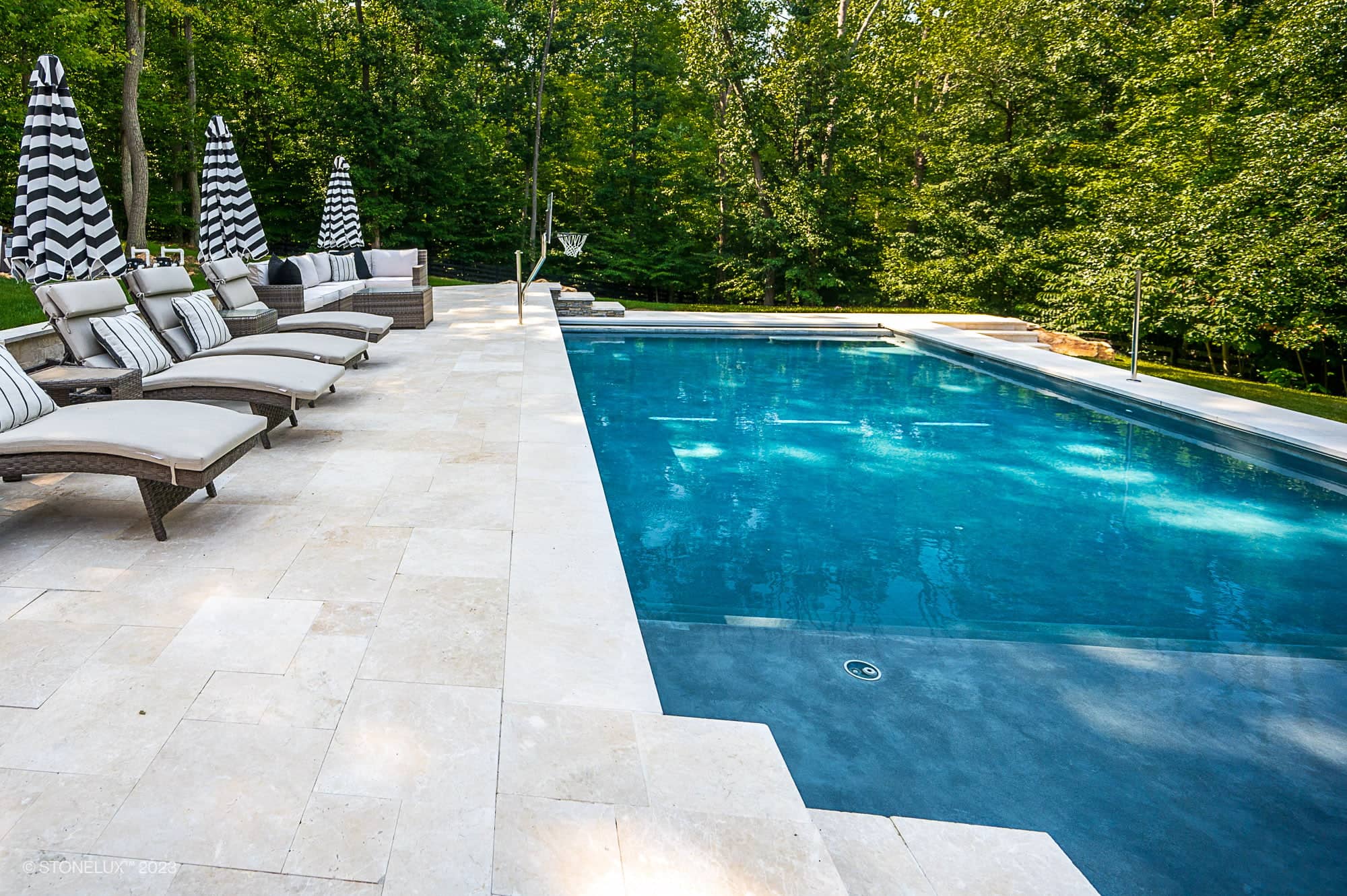 A modern outdoor swimming pool with lounge chairs featuring striped cushions and black-and-white umbrellas, set on a stone patio of Pera Cream Marble among the trees, bordered by lush greenery.