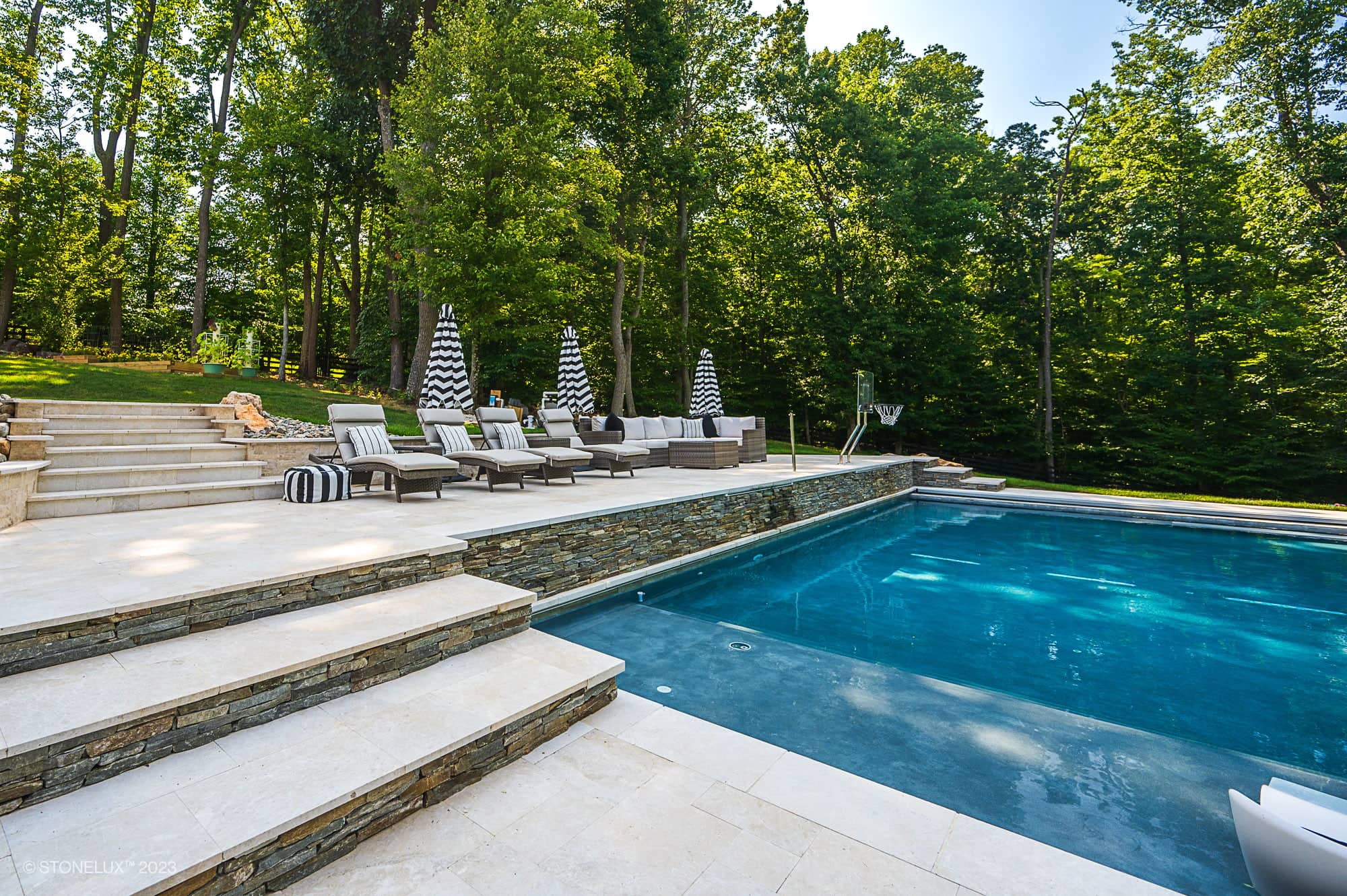 Luxurious outdoor pool area with Pera Cream Marble stone steps, lounge chairs, striped umbrellas, and lush green trees in the background on a sunny day.