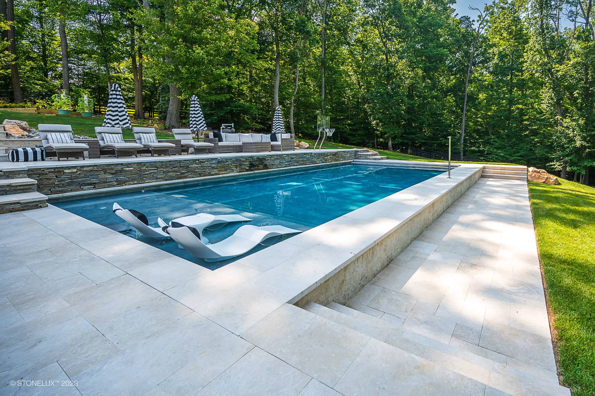 Modern outdoor swimming pool with two white lounge chairs partially submerged, surrounded by Pera Cream Marble decking, grassy lawn, and a seating area with striped umbrellas and cushioned sofas. Trees fill the background.