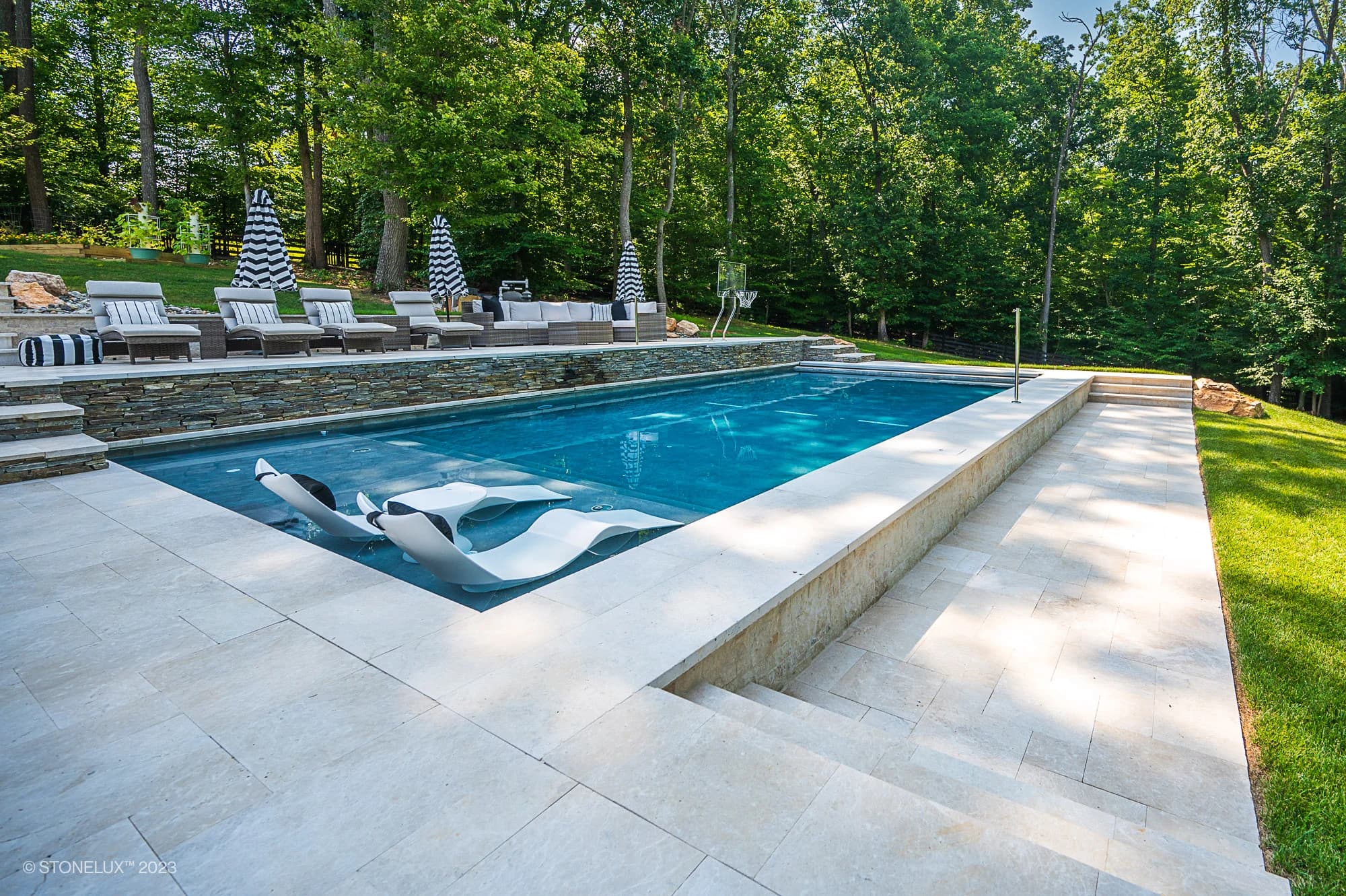 Modern outdoor swimming pool with two white lounge chairs partially submerged, surrounded by Pera Cream Marble decking, grassy lawn, and a seating area with striped umbrellas and cushioned sofas. Trees fill the background.